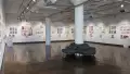 The first gallery space is shown. Three walls are shown, and display several groups of prints all throughout. Each group of prints has a personalized book under the prints on floating shelves. A pedestal sits near the back of the gallery space to the left, with a sculpture of three figures on top the hood of a car. Another pedestal sits near the front on the right, with a sculpture of three pizza boxes, stacked on top of eachother. Two large pillars rest in the middle of the gallery space. 