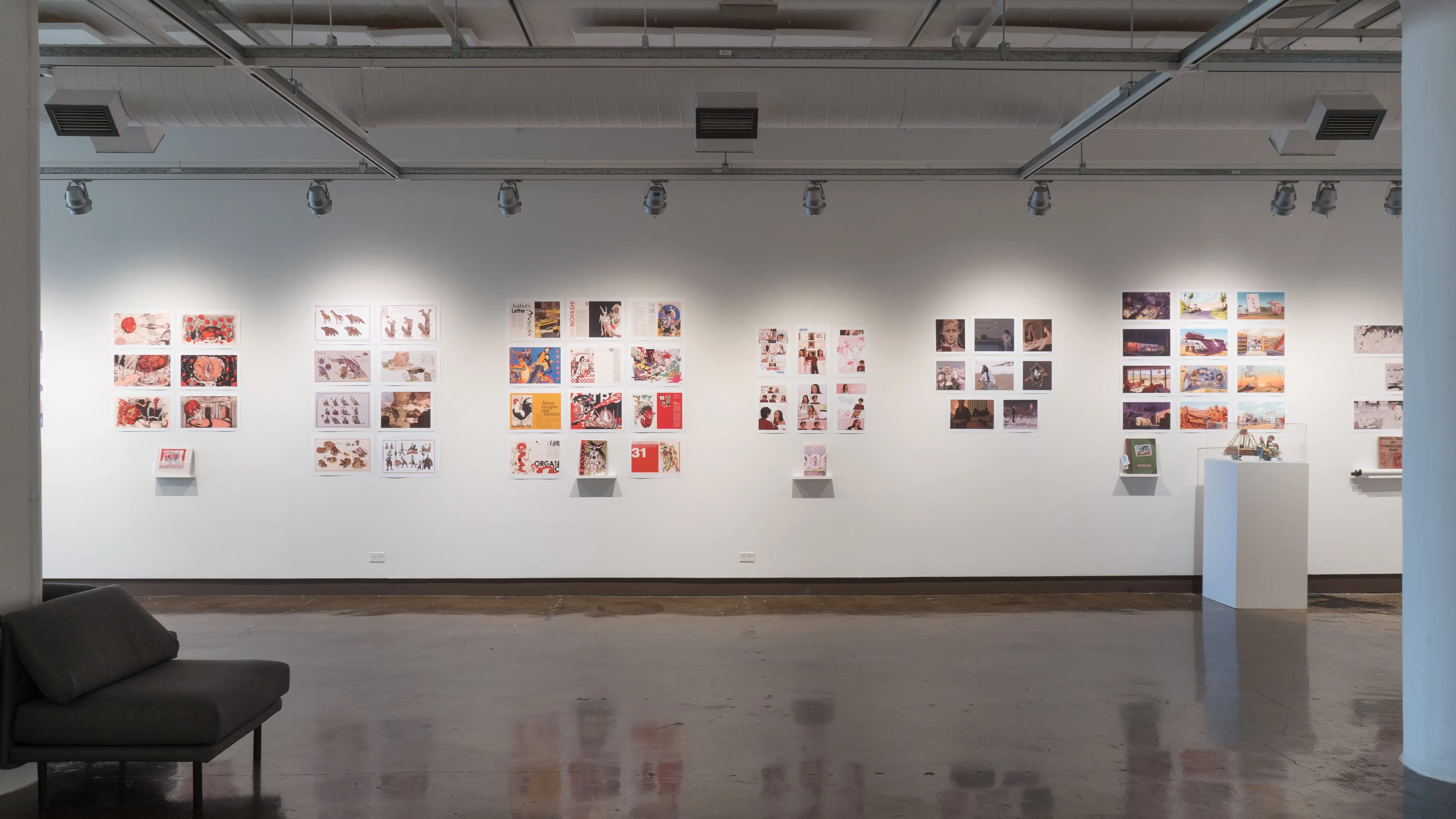 In the first gallery space, a wall is shown displaying seven groups of prints, with the last group of prints to the right cut off by a pillar. The first, third, fourth, sixth, and seventh group of prints all have personalized books displayed under the prints on floating shelves. The sixth group of prints has a pedestal with a sculpture of three figures on top of the hood of a car.