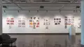 In the first gallery space, a wall is shown displaying seven groups of prints, with the last group of prints to the right cut off by a pillar. The first, third, fourth, sixth, and seventh group of prints all have personalized books displayed under the prints on floating shelves. The sixth group of prints has a pedestal with a sculpture of three figures on top of the hood of a car.