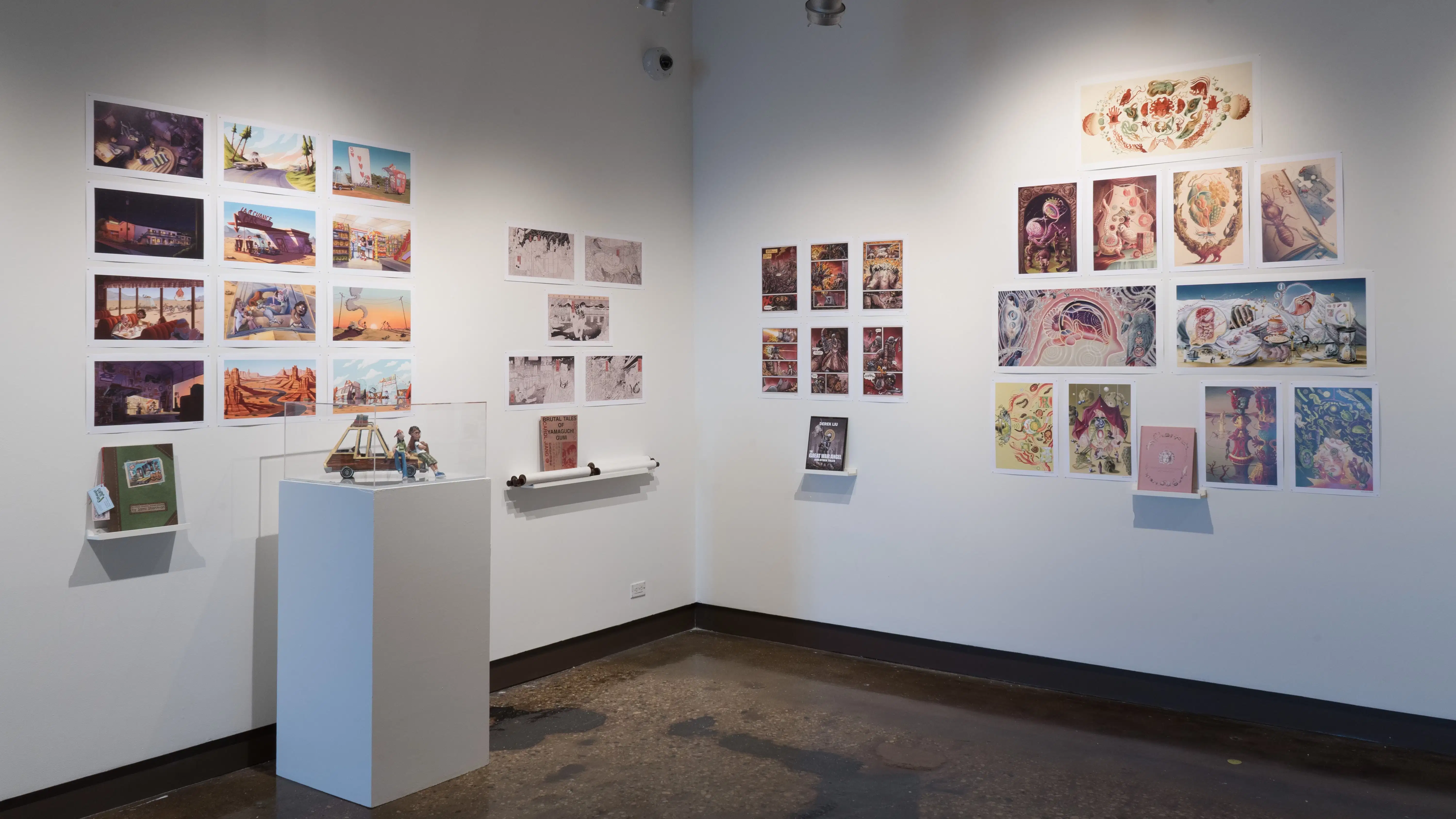 In the corner of the first gallery space, four groups of vertical and horizontal prints are displayed. Each group of prints has a personalized book displayed under the prints on floating shelves. On the left wall, the first set of prints has a pedestal with a sculpture of three figures on top of the hood of a car. The second group of prints has several scrolls holding up the personalized book.