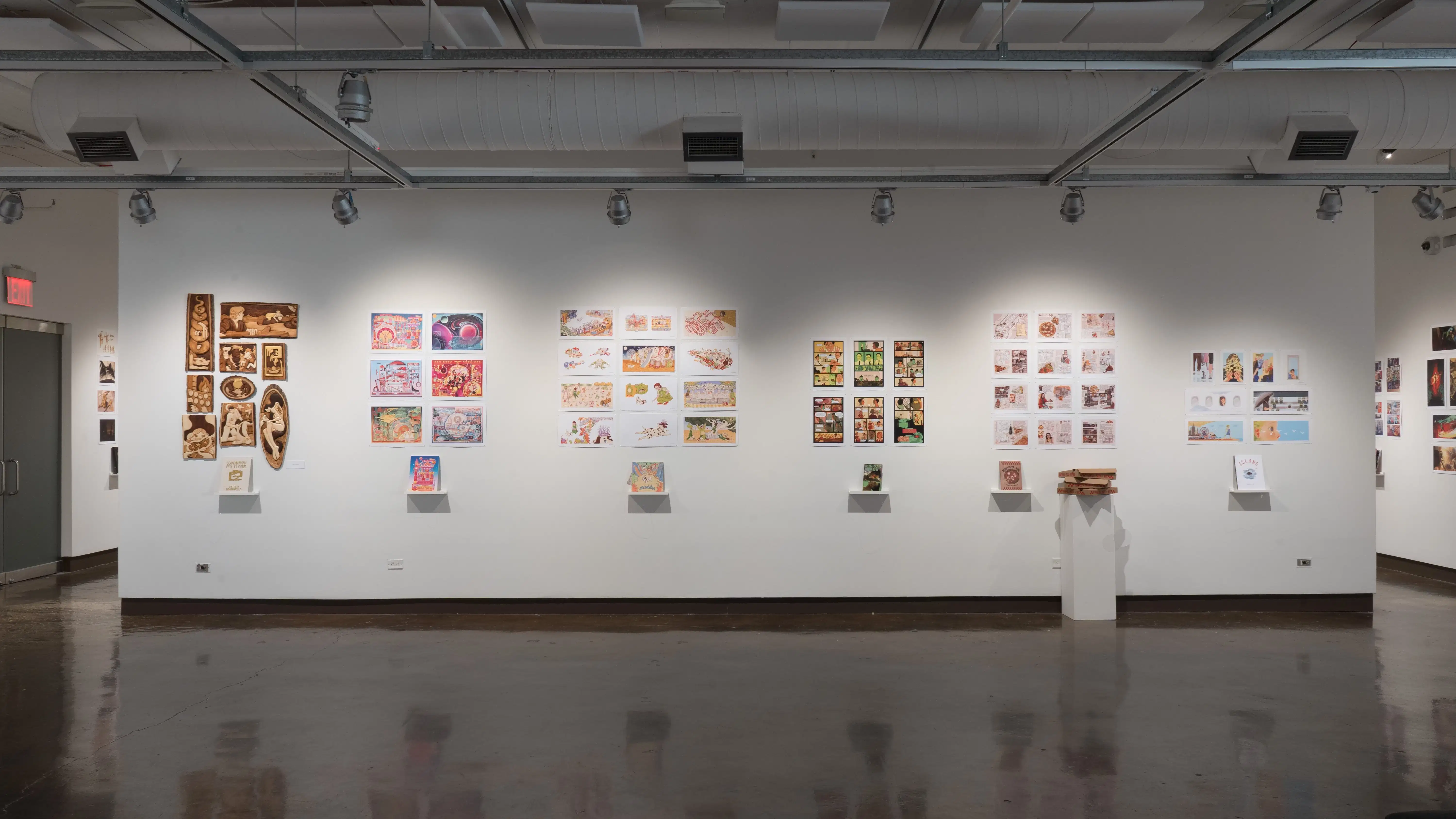 In the first gallery space, six groups of prints are displayed, with each group having a personalized book displayed under the prints on floating shelves. The first group of prints on the left hand side is made out of carved wood. The fifth group is accompanied with a sculpture of three pizza boxes on a pedestal. The rest of the prints are all on paper.
