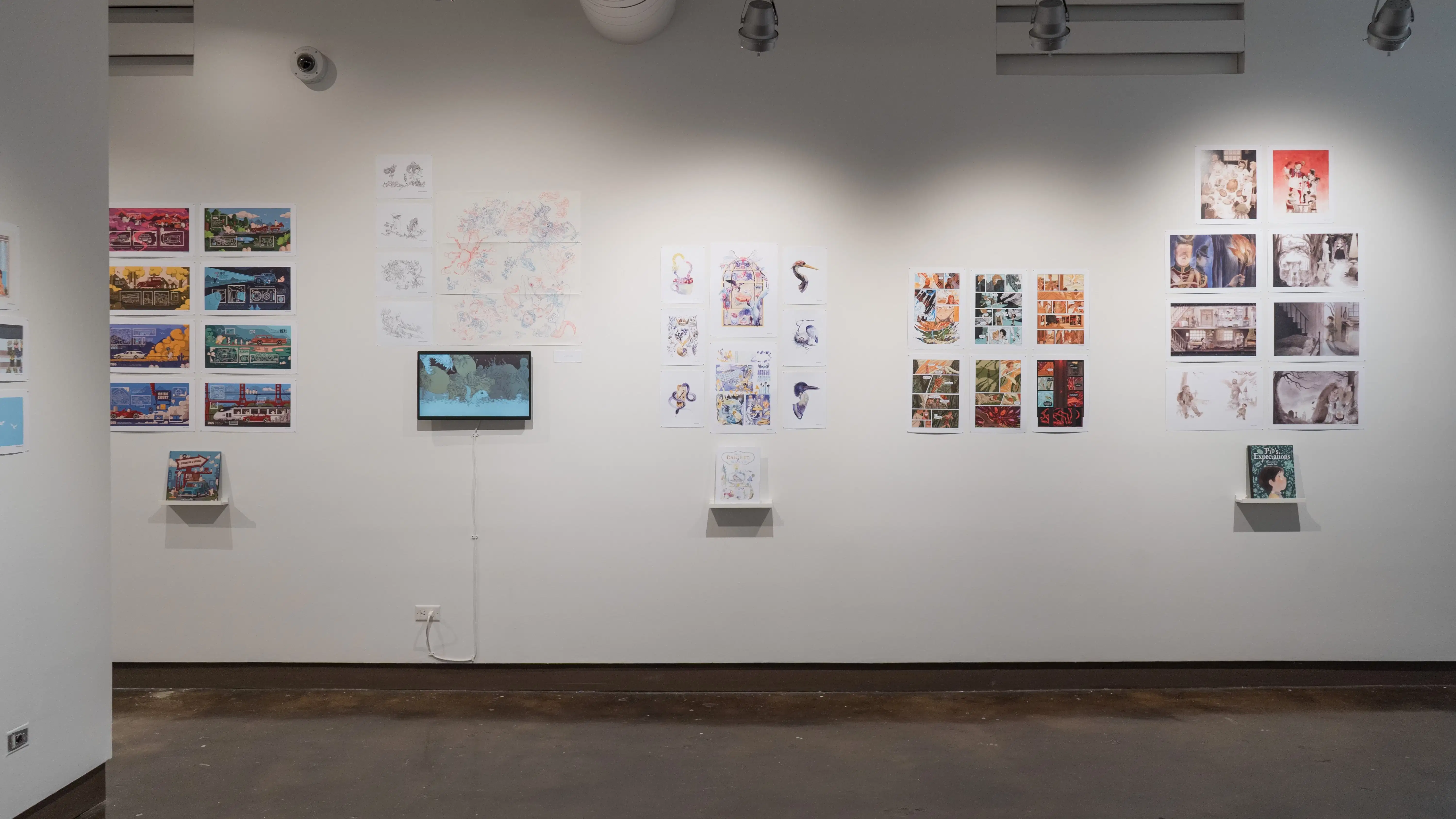 In the first gallery space, five groups of horizontal and vertical prints are shown. From left to right, the first, third, and fifth group of prints all have books displayed under the prints on floating shelves. The second group of prints is accompanied with a monitor displaying an animation.