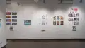 In the first gallery space, five groups of horizontal and vertical prints are shown. From left to right, the first, third, and fifth group of prints all have books displayed under the prints on floating shelves. The second group of prints is accompanied with a monitor displaying an animation.