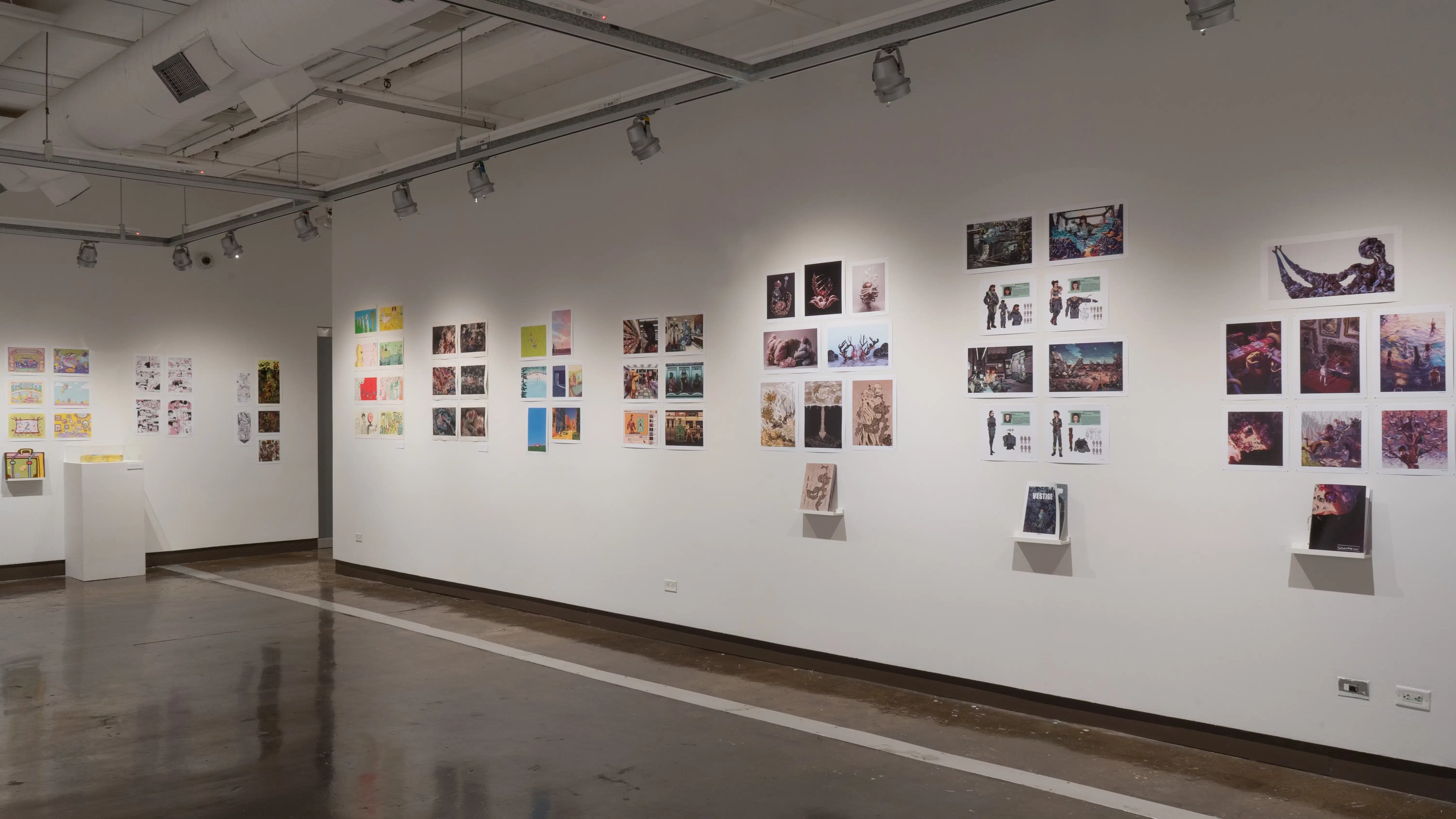 In the second gallery space, two walls are shown. On the left, a wall displays three groups of several prints. The first group of prints has a personalized book displayed on a floating shelf under the prints, and a sculpture on a pedestal. On the right, seven groups of prints are displayed, with the last three nearest to the right having a personalized book displayed under the prints on separate floating shelves.
