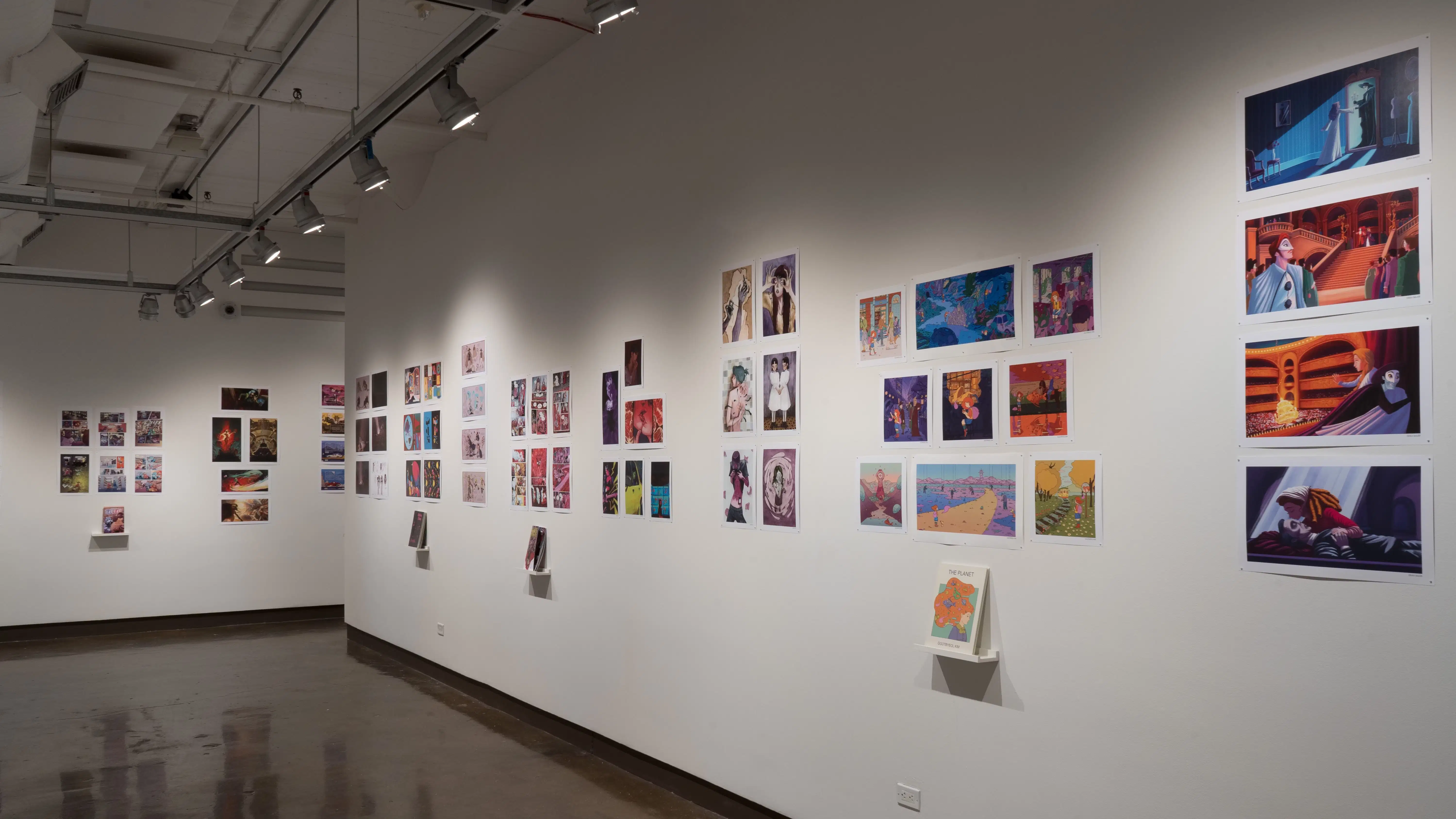 In the second gallery space, two walls are shown. The wall furthest shown displays several horizontal and vertical printed works, some accompanied with a book displayed on a floating shelf under the prints. To the right, a wall displays several horizontal and vertical prints. All the prints range in size and some grouped prints have a personalized book displayed under the prints on floating shelves.