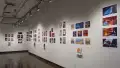 In the second gallery space, two walls are shown. The wall furthest shown displays several horizontal and vertical printed works, some accompanied with a book displayed on a floating shelf under the prints. To the right, a wall displays several horizontal and vertical prints. All the prints range in size and some grouped prints have a personalized book displayed under the prints on floating shelves. 