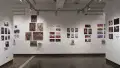 In the second gallery space, three walls are shown. Each wall consists of several horizontal and vertical prints. Some of these prints are accompanied with a personalized book displayed on floating shelves under the prints. On the center wall to the left, there are 6 paintings displayed. 