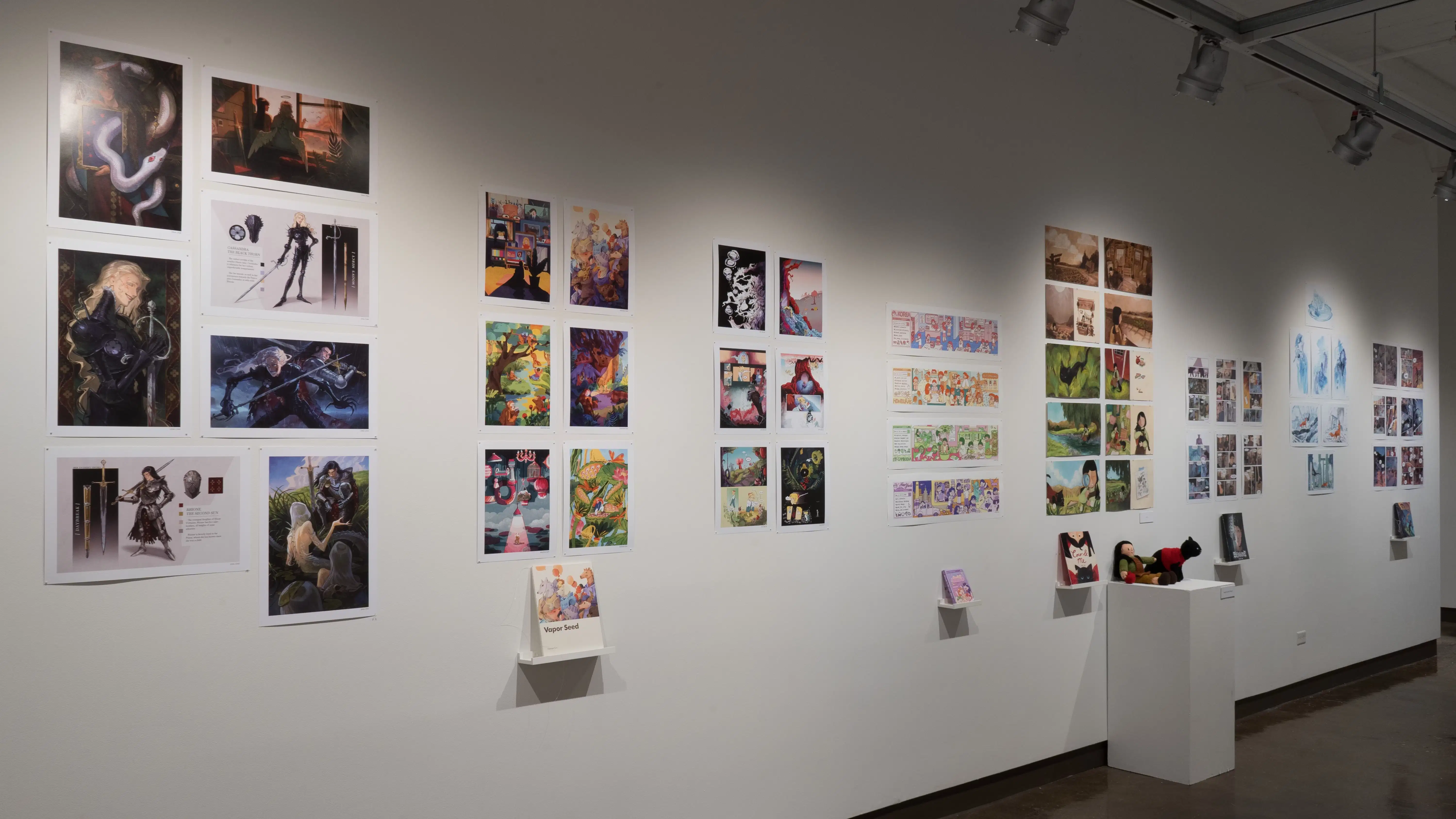In the first half of the last room of the gallery, several horizontal and vertical prints are shown on the wall. Some of these grouped prints are accompanied with a personalized book. In the center of the wall, a pedestal displays two crocheted figures; a little girl and a black cat.