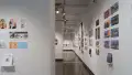 In the first room of the gallery, a hallway is shown, with many walls displaying several horizontal and vertical prints to the left and right. On the left, there are 7 prints; the top three are vertical, and the bottom four are horizontal. A book is displayed on a floating shelf under these prints. To the left and down the hallway, several groups of prints are displayed, some accompanied with a book displayed under the prints. The furthest wall to the back displays several books on two long floating shelves.