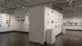 In the first half of the last room of the gallery, four walls are shown. In the room on the left, there are two walls that display several vertical and horizontal prints, some accompanied with personalized books. In the middle of all these prints, a monitor displays an animation. Around the corner, another wall displays several horizontal and vertical prints, some accompanied with a book displayed under the prints. Also shown under these prints are two pedestals on either side of the wall. One pedestal holds a book, and the other holds a dog figurine inside a box. In the wall furthest to the back on the right, two long floating shelves display several books, some accompanied with business cards.