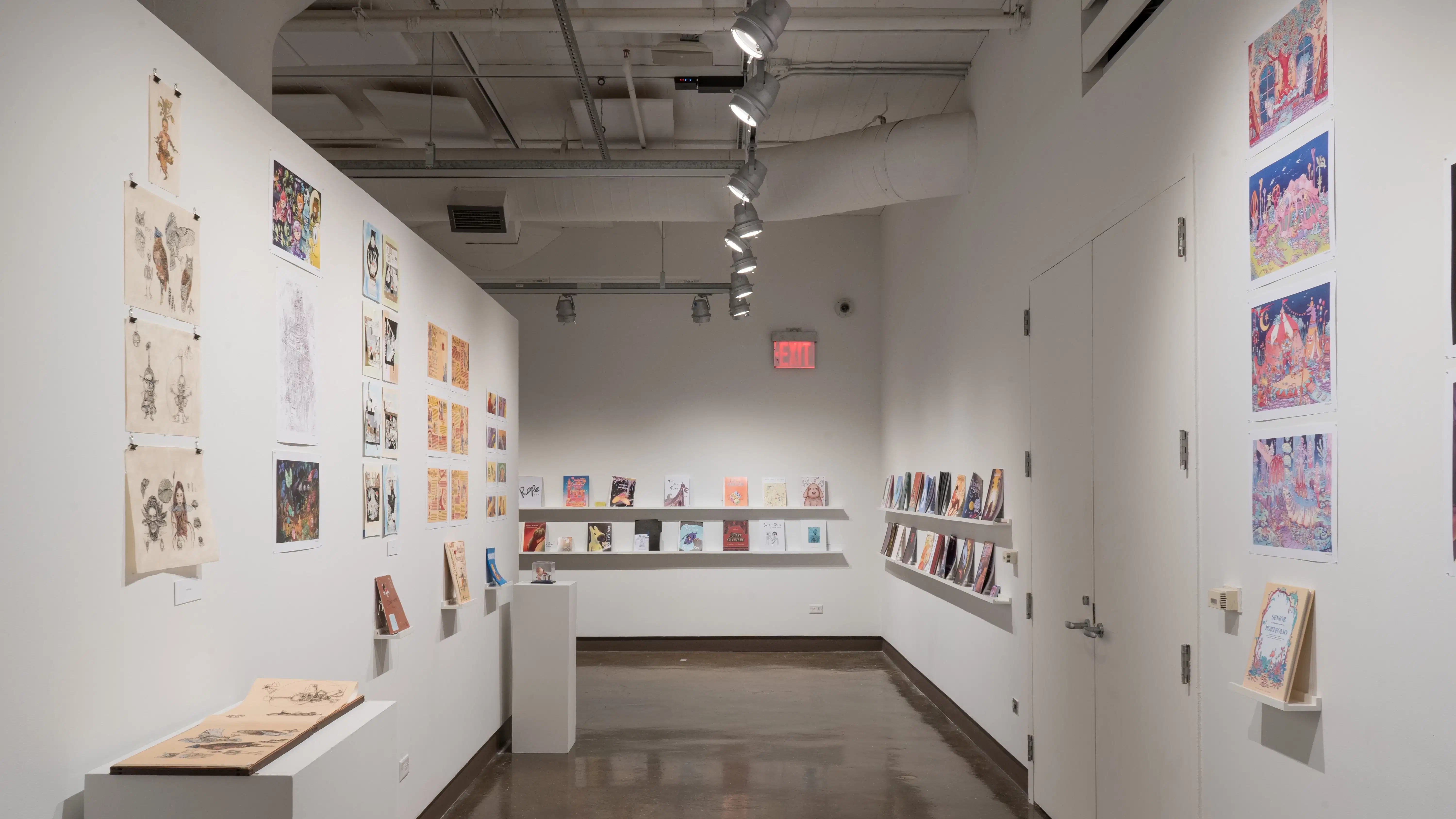 Down the hallway of the last space in the gallery, several printed works are displayed on the wall. On the left wall, several horizontal and vertical prints are displayed on the wall, most accompanied with a book displayed under the prints on floating shelves and/or a pedestal. The last group of prints has a dog figurine in a box displayed on a pedestal below the prints. The wall furthest shown displays several books on two long floating shelves, some accompanied with business cards. On the wall to the right, several books are displayed on two long floating shelves, some accompanied with business cards. To the edge of the image shown, four horizontal prints are displayed with a personalized book displayed under the prints on a floating shelf.