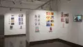 In the first half of the last room of the gallery, there are three walls shown. On the right wall, four prints are displayed. To the right of the prints, a screen displays an animation. On the center wall to the left of the right wall, two groups of prints are displayed with personalized books displayed under the prints on floating shelves. The furthest wall around the corner and behind the main two walls shown displays several horizontal and vertical prints, with books displayed under the two groups of prints.