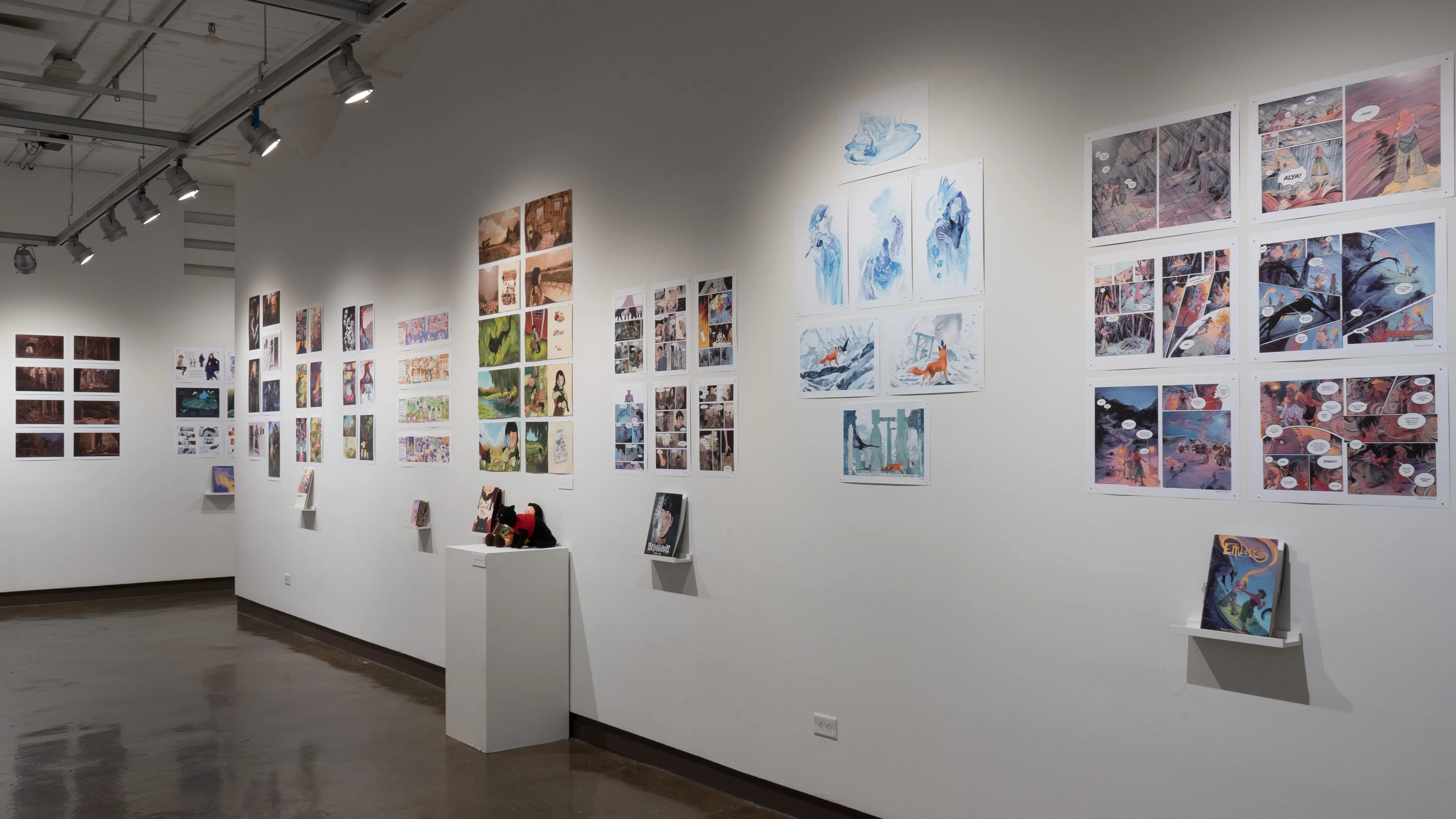 In the first half of the last room of the gallery, there are two walls photographed. On the furthest wall to the left, 8 horizontal prints are displayed, and more prints are displayed, but are cut off by the main wall photographed. The main wall to the right displays several horizontal and vertical prints, some accompanied with a book. In the center of the wall, there are two crocheted figures; one is a black cat and the other is of a little girl.