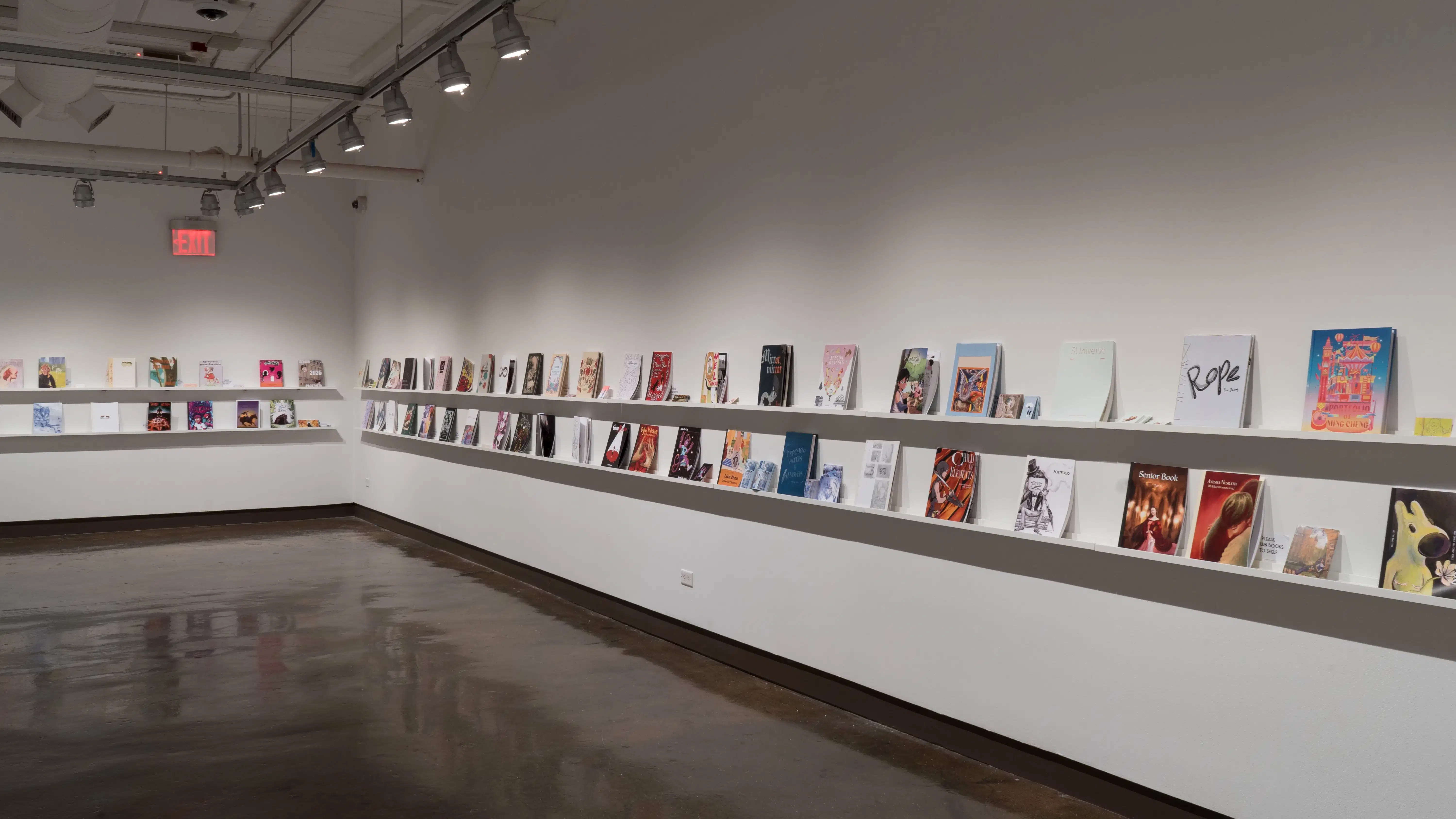 In the last room of the gallery, two walls are shown, with two long shelves displaying several books. The furthest wall to the left displays 13 books, some accompanied with business cards. The wall to the right displays 44 books, some accompanied with business cards and/or stickers.