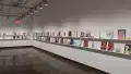In the last room of the gallery, two walls are shown, with two long shelves displaying several books. The furthest wall to the left displays 13 books, some accompanied with business cards. The wall to the right displays 44 books, some accompanied with business cards and/or stickers.