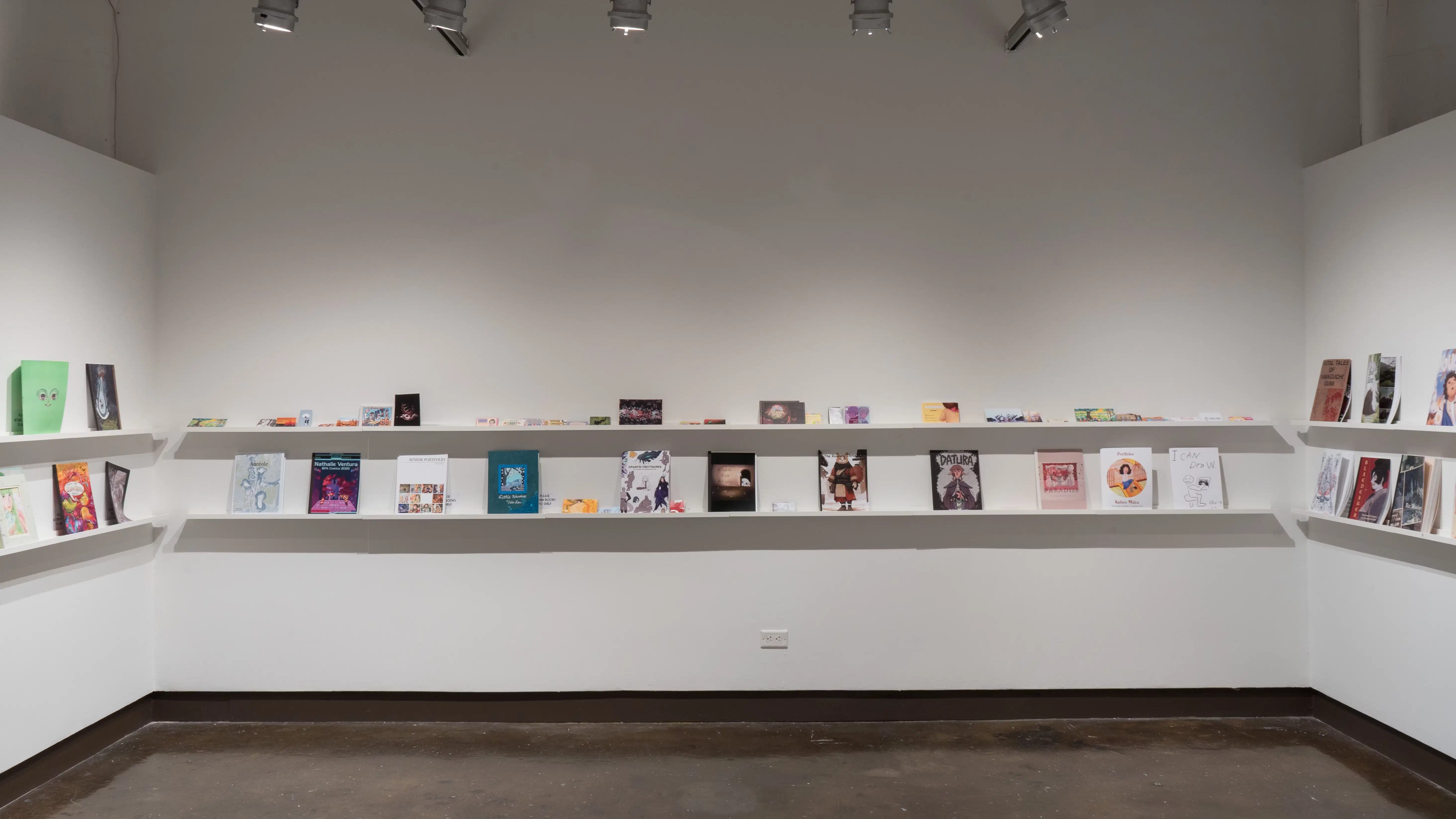 In the last room of the gallery, there are three walls that have several books displayed on two long shelves. On the left wall, five books are displayed. On the center wall, the top shelf holds several artists' business cards, and the bottom shelf holds books and business cards. The right wall displays 6 books.