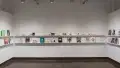 In the last room of the gallery, there are three walls that have several books displayed on two long shelves. On the left wall, five books are displayed. On the center wall, the top shelf holds several artists' business cards, and the bottom shelf holds books and business cards. The right wall displays 6 books.