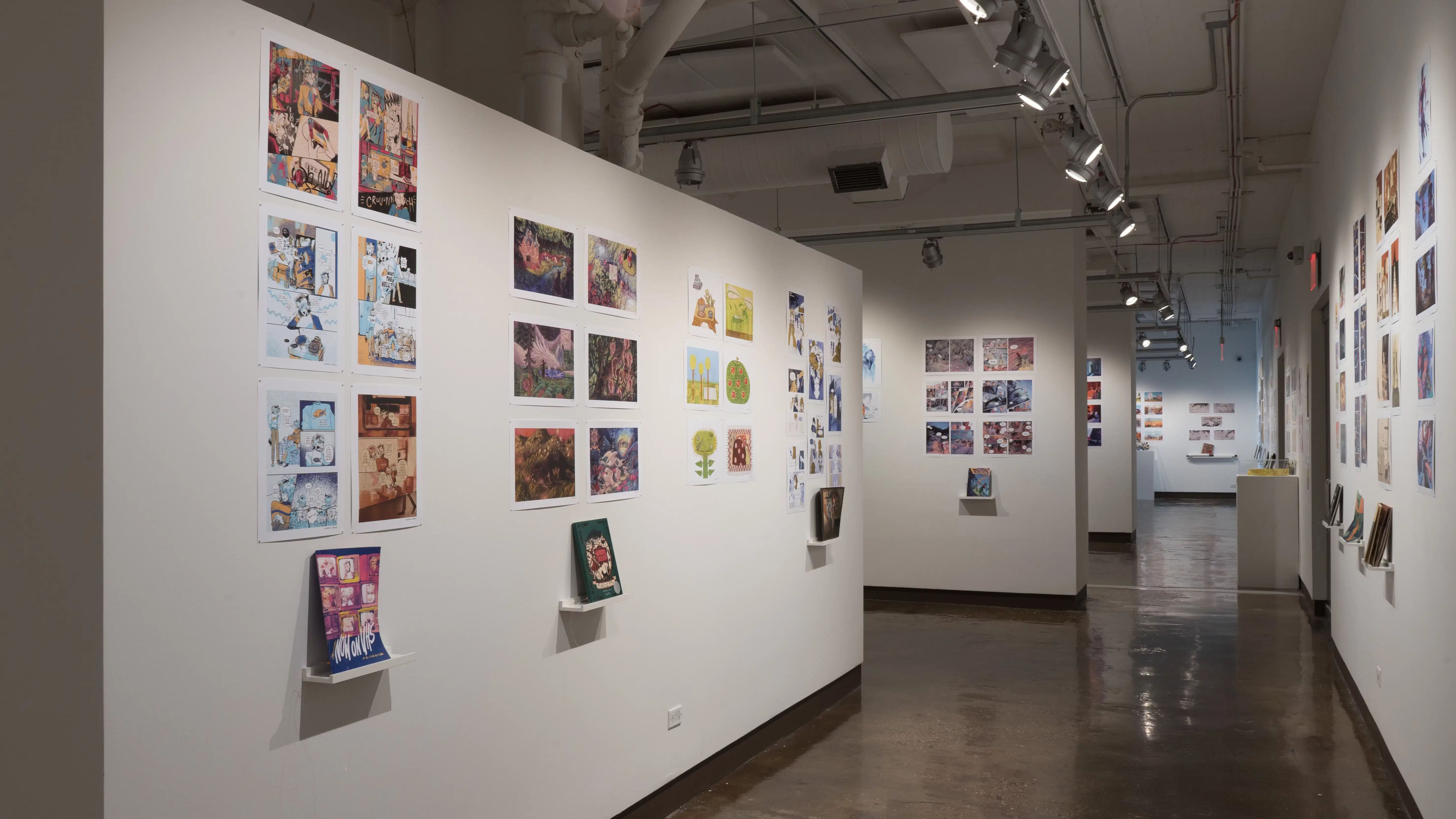 Three walls are shown, with a corridor down the middle. On the left wall, four different artists' works are displayed, all consisting of prints similar in size. Three of the works are accompanied with a book on a shelf. The first three works consist of 6 prints each, while the last work consists of 8 prints.