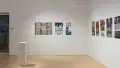 A stright shot of the SVA Flatiron Gallery's south and west walls, showing many colorful prints by BFA Animation students pinned up; some paper sits atop a pedestal in the left foreground. The glass doors are visible in the back left.