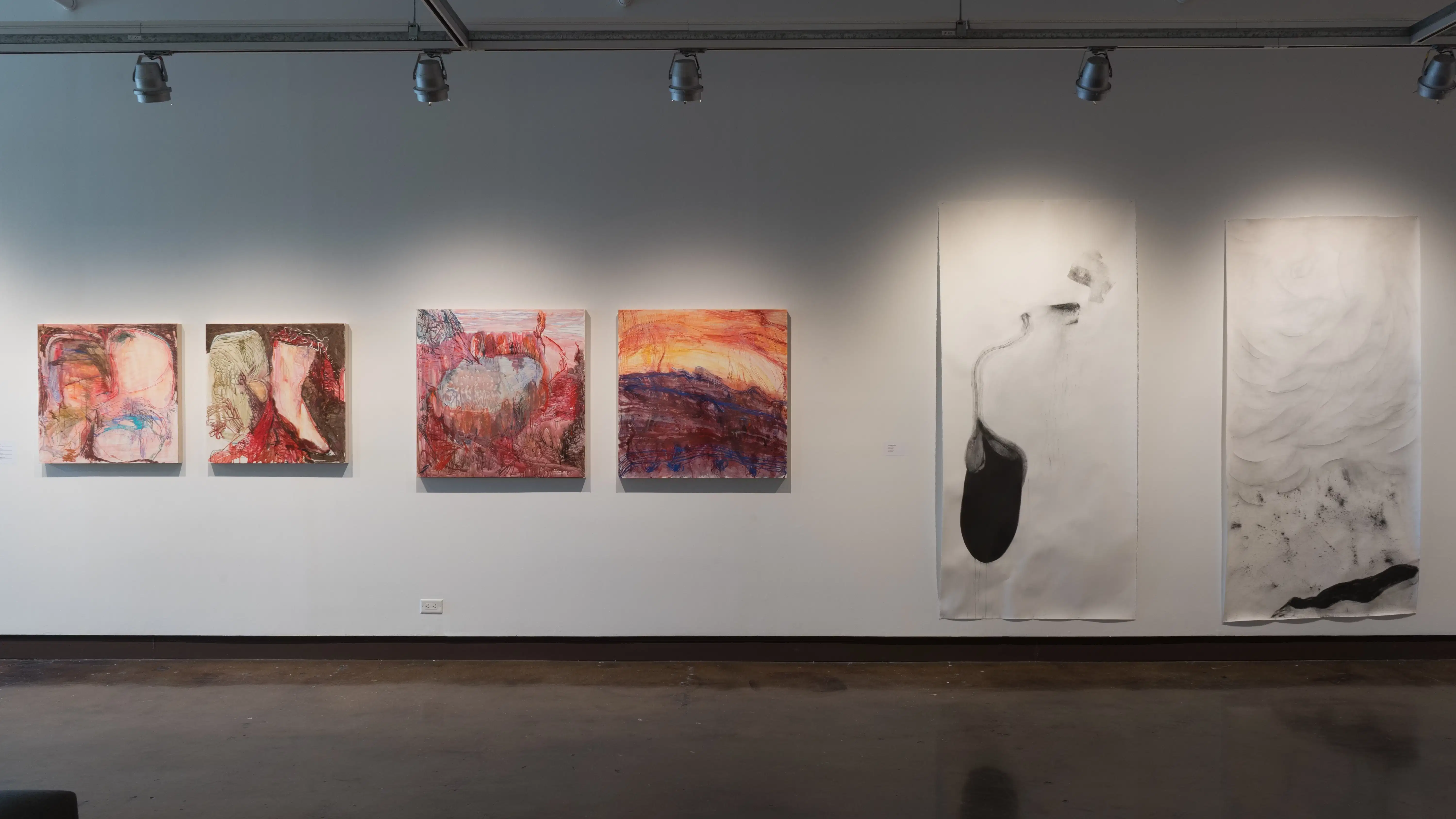Along one of the walls in gallery one, four square, colorful abstract paintings are hung in a row. Beside them are two charcoal drawings on long, vertical, mostly white pieces of paper.