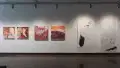 Along one of the walls in gallery one, four square, colorful abstract paintings are hung in a row. Beside them are two charcoal drawings on long, vertical, mostly white pieces of paper.