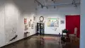 A room-like installation is set up in the corner of the space, with a large painting, a desk, a candle, a bookshelf, and other objects. To the left of the installation, a charcoal drawing on a long, vertical, mostly white piece of paper is hung on the wall.