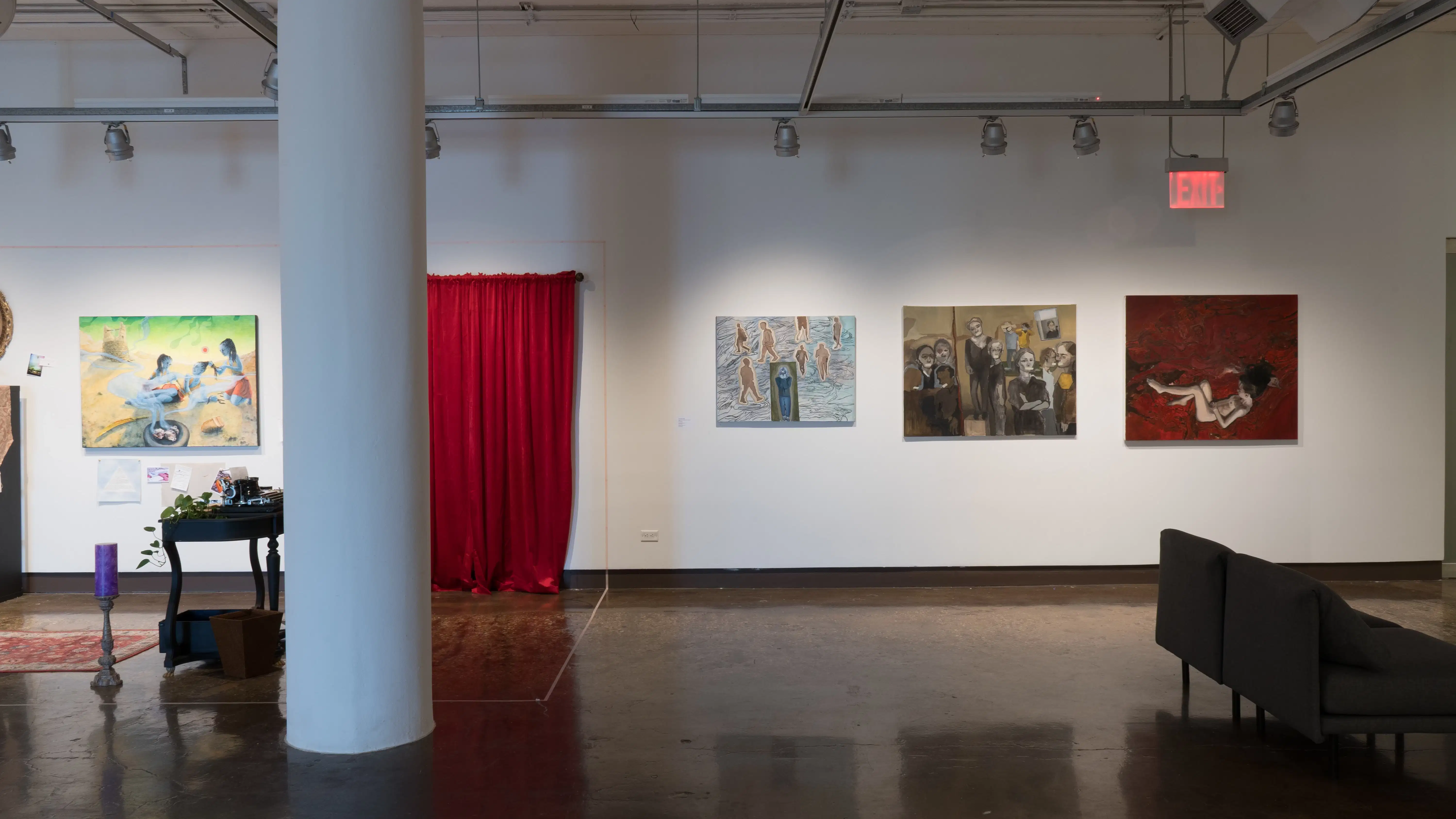 On the left, a room-like installation is set up in the corner of the space, with a large painting, a desk, a candle, and a red velvet curtain. To the right of this installation, three paintings on canvas are hung in a row.