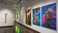 On the right wall of gallery one, three large paintings depict human figures merging with nature in various states. One painting is monotone sepia, while the other two are a vibrant cobalt with pinks and yellows. A mixed media assemblage is visible on the wall to the left.