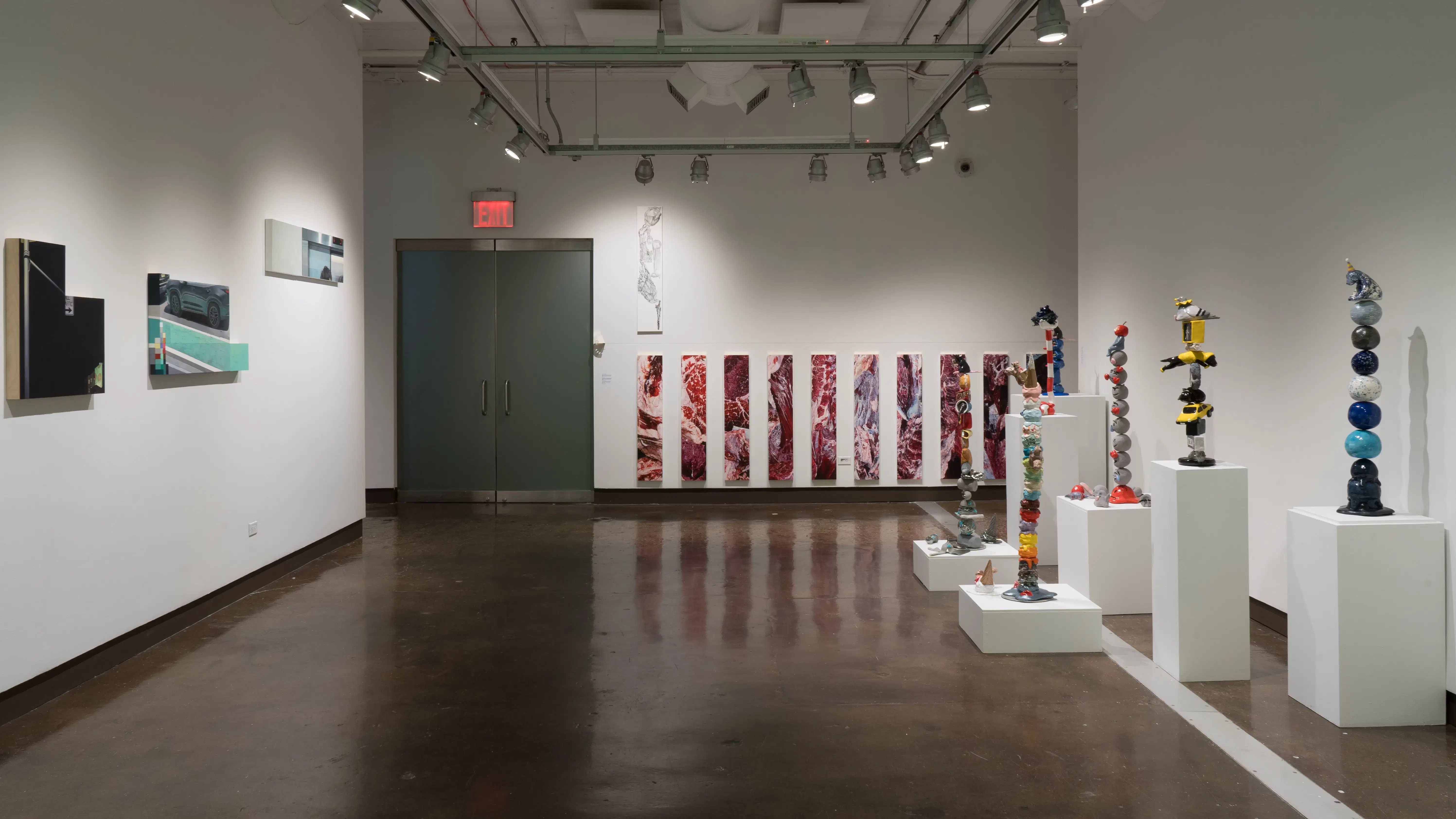 On the far wall in gallery two, narrow, vertical paintings that look like raw meat are hung in a row. On the left wall are the paintings that seem to be cut from a whole, and on the right are several pedestals displaying ceramic sculptures of objects stacked precariously high.