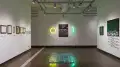 On the far wall in gallery two, a painting of pigeons in a space-like environment hangs between two neon lights. The yellow and green lights reflect on blue, textured resin tiles that have been arranged on the floor. Framed works hang on the left wall, and canvas paintings hang on the right.