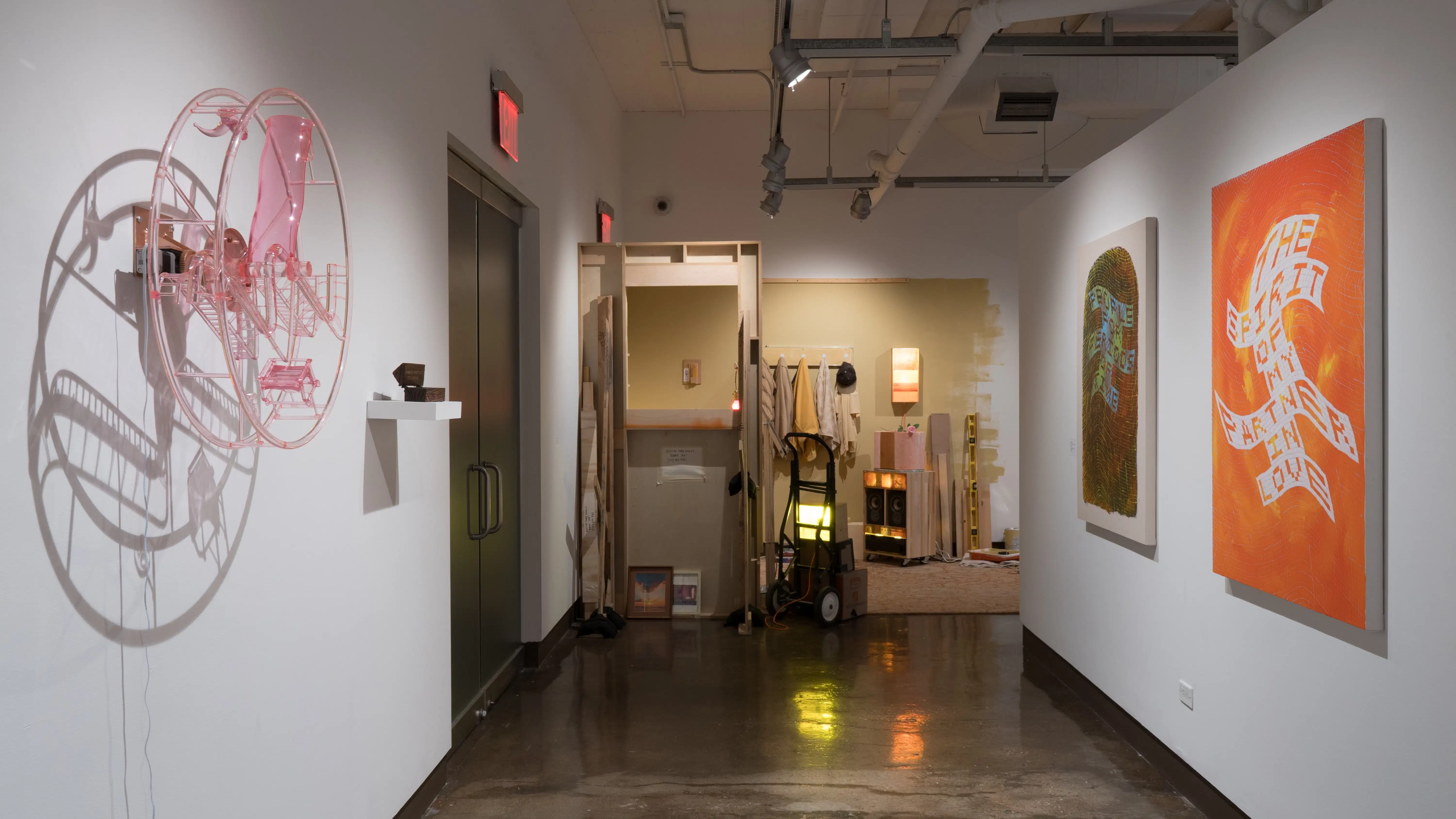 In the left hallway of the gallery, a pink acrylic wheel is mounted on the wall beside a small wooden box on a shelf. At the end of the hallway is a corner installation of a room-like space. On the right side of the hallway are two paintings on canvas using warped, blocky text.
