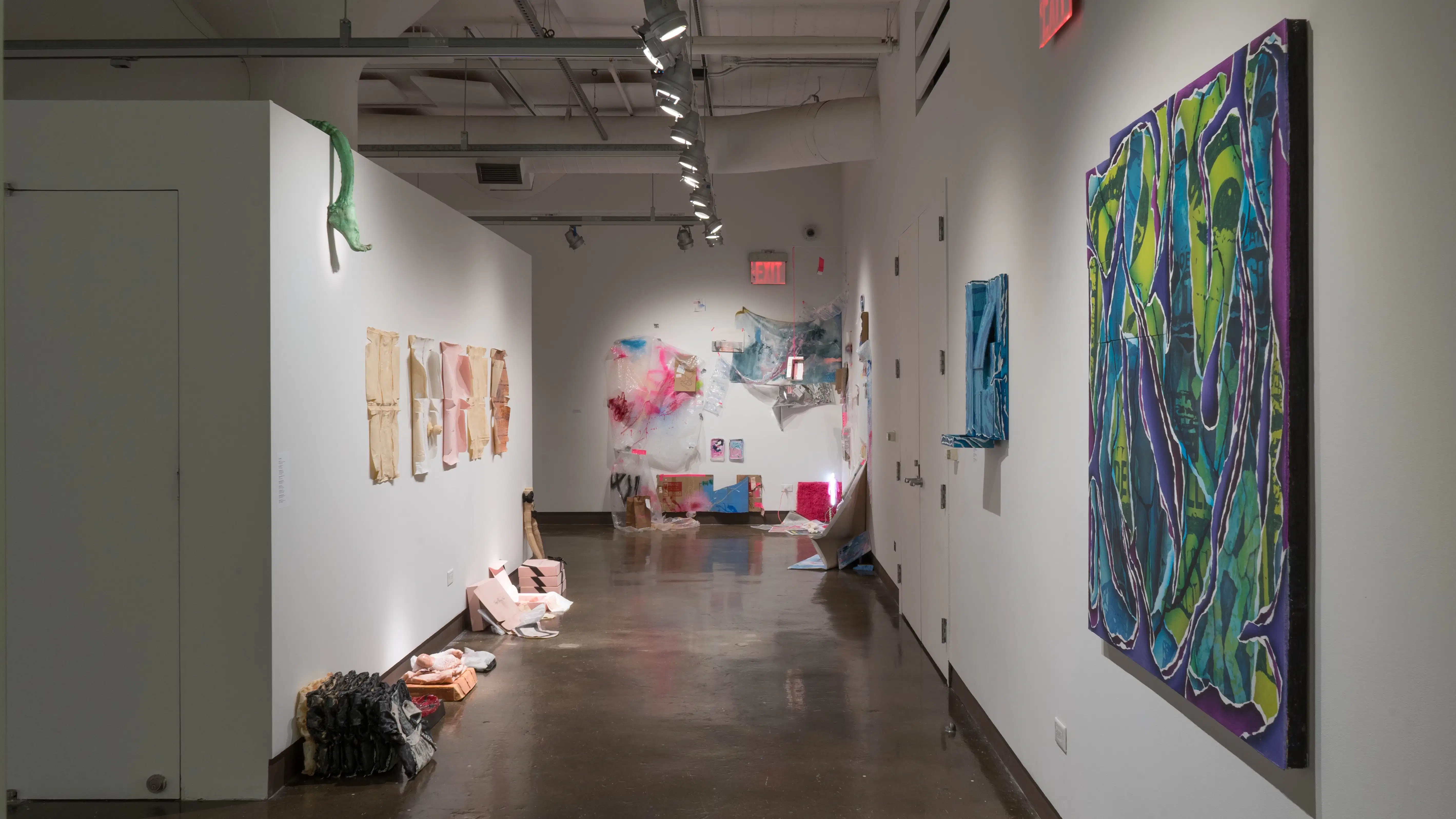 Facing the back of the gallery, paper works seemingly peel off of the left wall in the right hallway. Below them, several sculptures sit in a line on the floor. Paintings using blues, purples, and greens hang on the right side of the hall, and an installation is visible in the back corner.