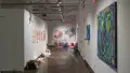 Facing the back of the gallery, paper works seemingly peel off of the left wall in the right hallway. Below them, several sculptures sit in a line on the floor. Paintings using blues, purples, and greens hang on the right side of the hall, and an installation is visible in the back corner.