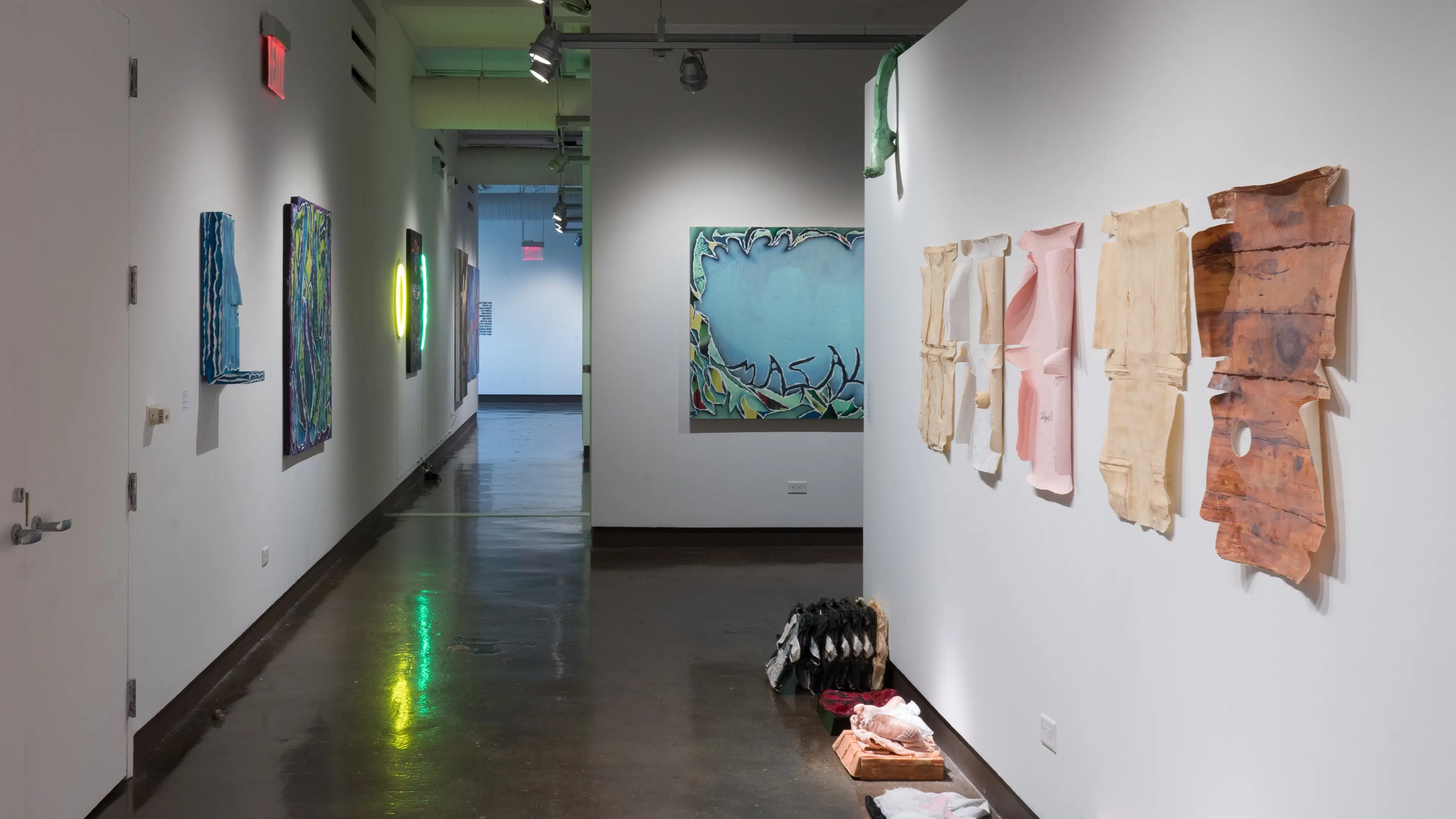 Looking back toward gallery one through the right hallway, some paper works displayed to the immediate right are seemingly peeling off the wall. Sculptures sit on the ground beneath them, while large paintings are visible on the left wall and in gallery 3 beyond them.