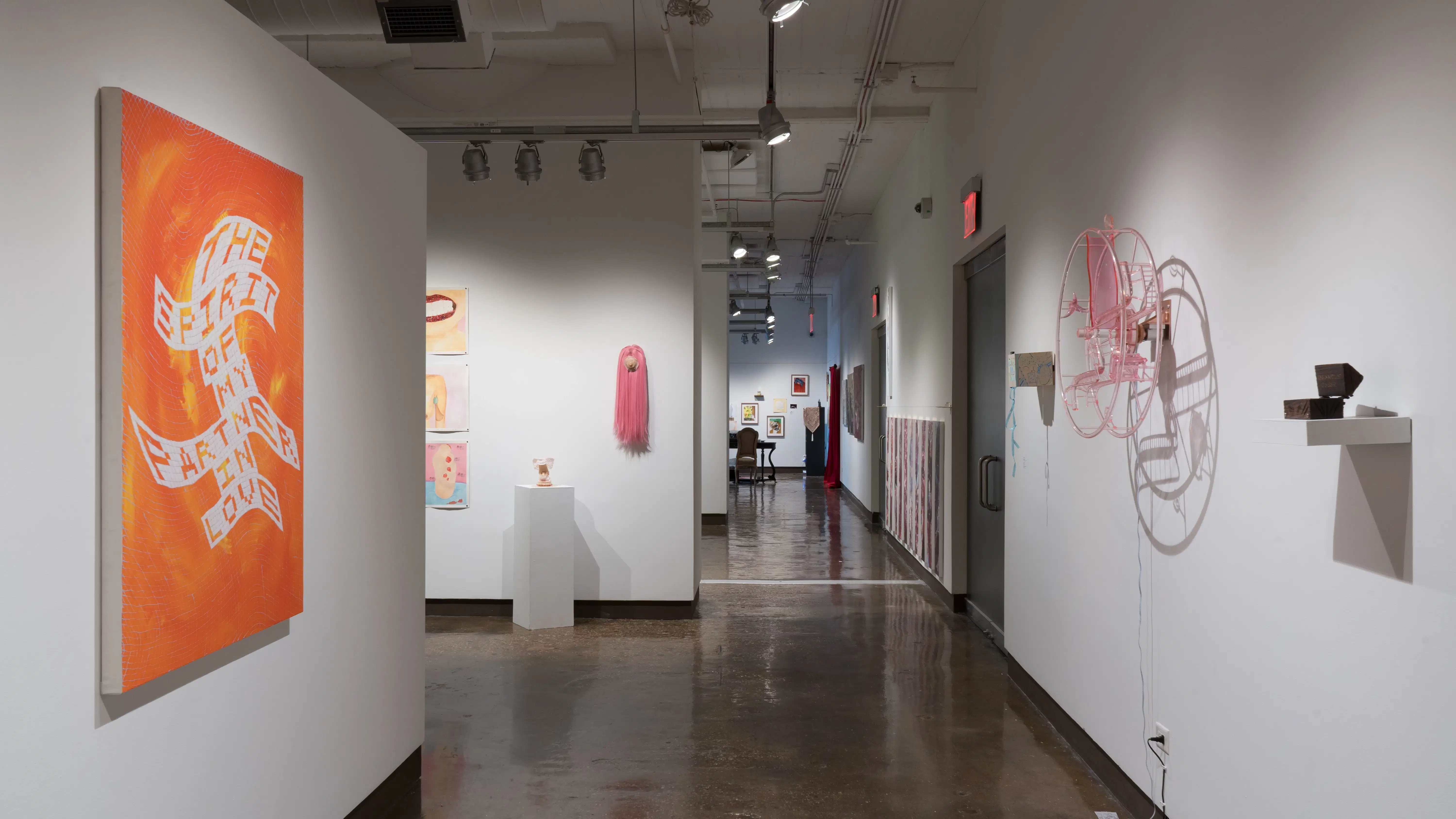 Looking back toward gallery one through the left hallway, an orange painting featuring blocky text is hung to the immediate left. To the right, a pink acrylic wheel is mounted on the wall beside a small wooden box. More works are visible in galleries three, two, and one.