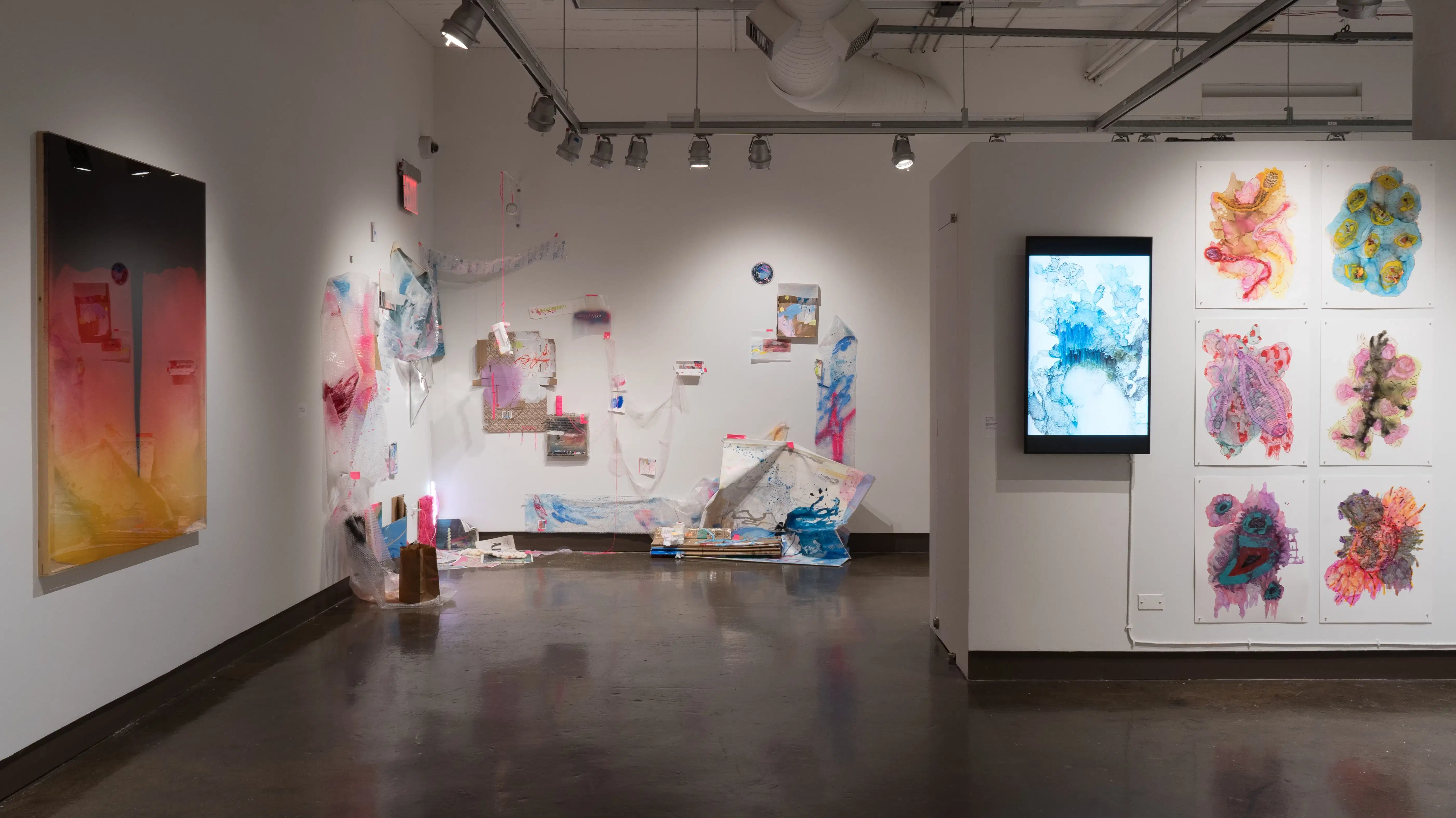 One half of gallery four is shown, with the pink and blue detritus installation, the bright watercolors and monitor, and one cat-eye-like painting all in view.