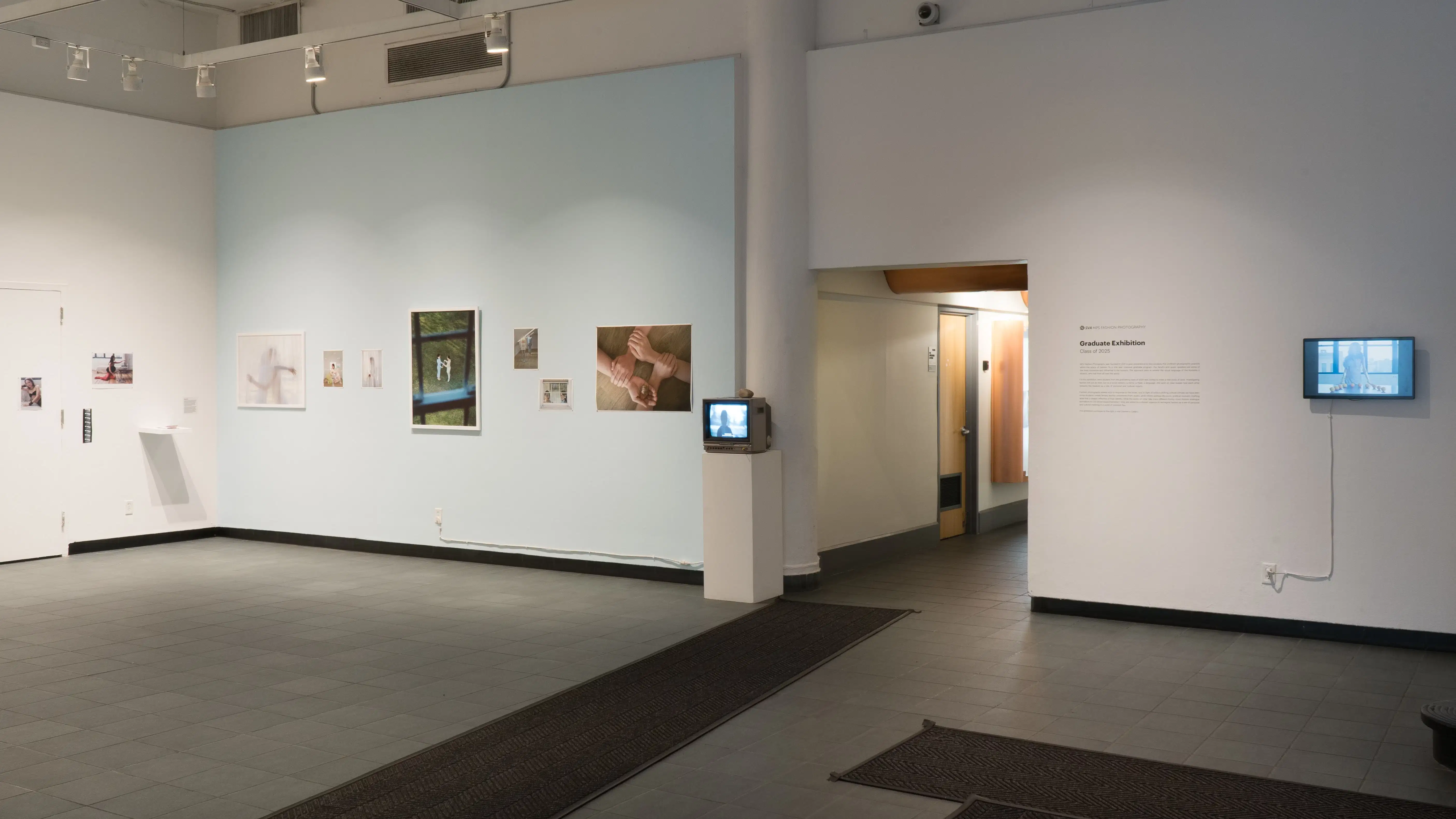 The view from the entrance into the SVA Gramercy Gallery. Two screens display video art, and a light blue wall to the left has a series of photographs on it. There are more photographs on the wall perpendicular to the light blue one.