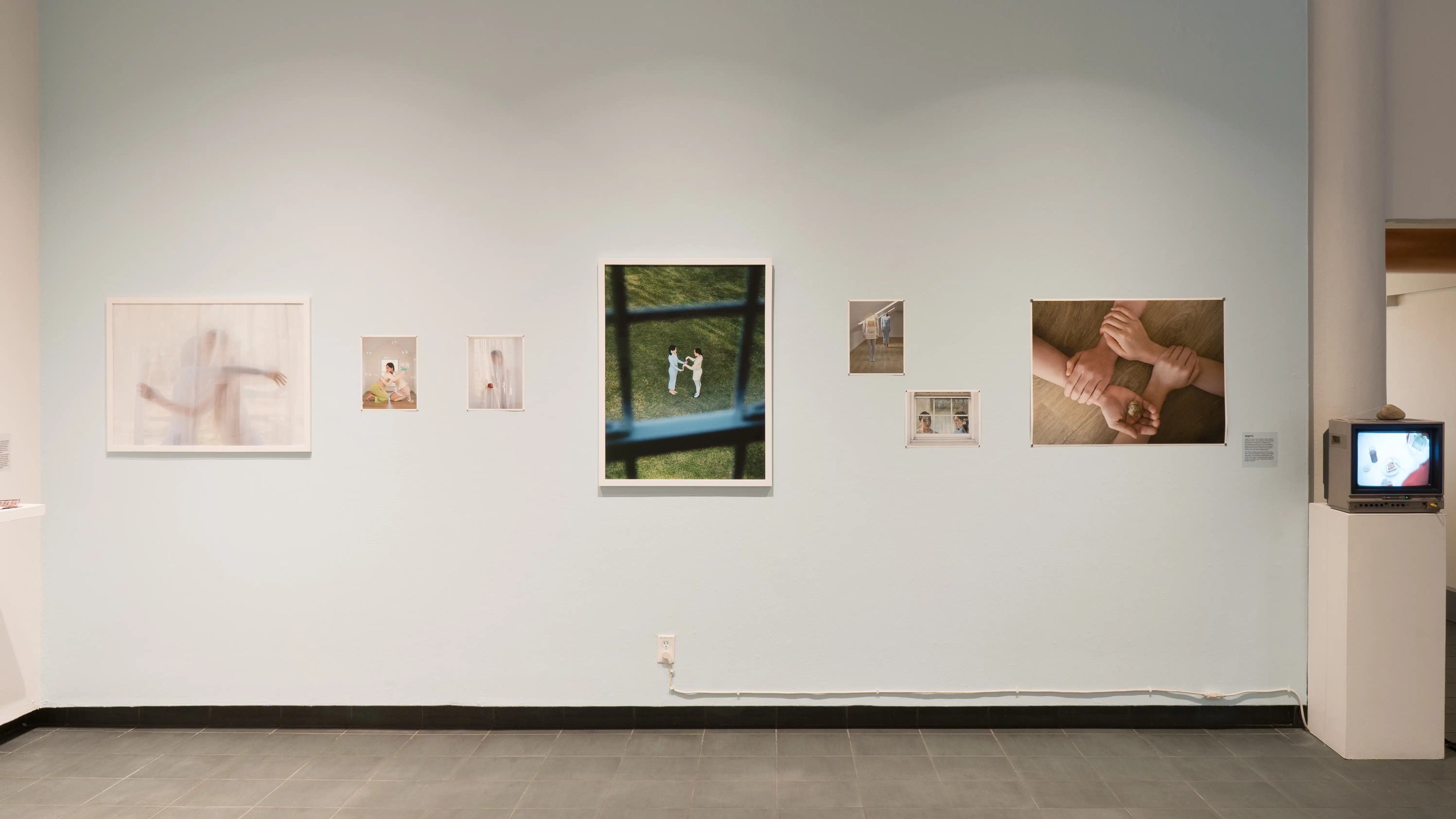 A series of dreamlike images on a light blue wall in the gallery lobby. To the right of the photographs is a small box television playing a video.