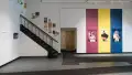 The lobby of the SVA Gramercy Gallery featuring work by a student. Three large vertical stripes of pink, yellow, and blue are painted on the wall. Photographs of a model posing are hung on these stripes.