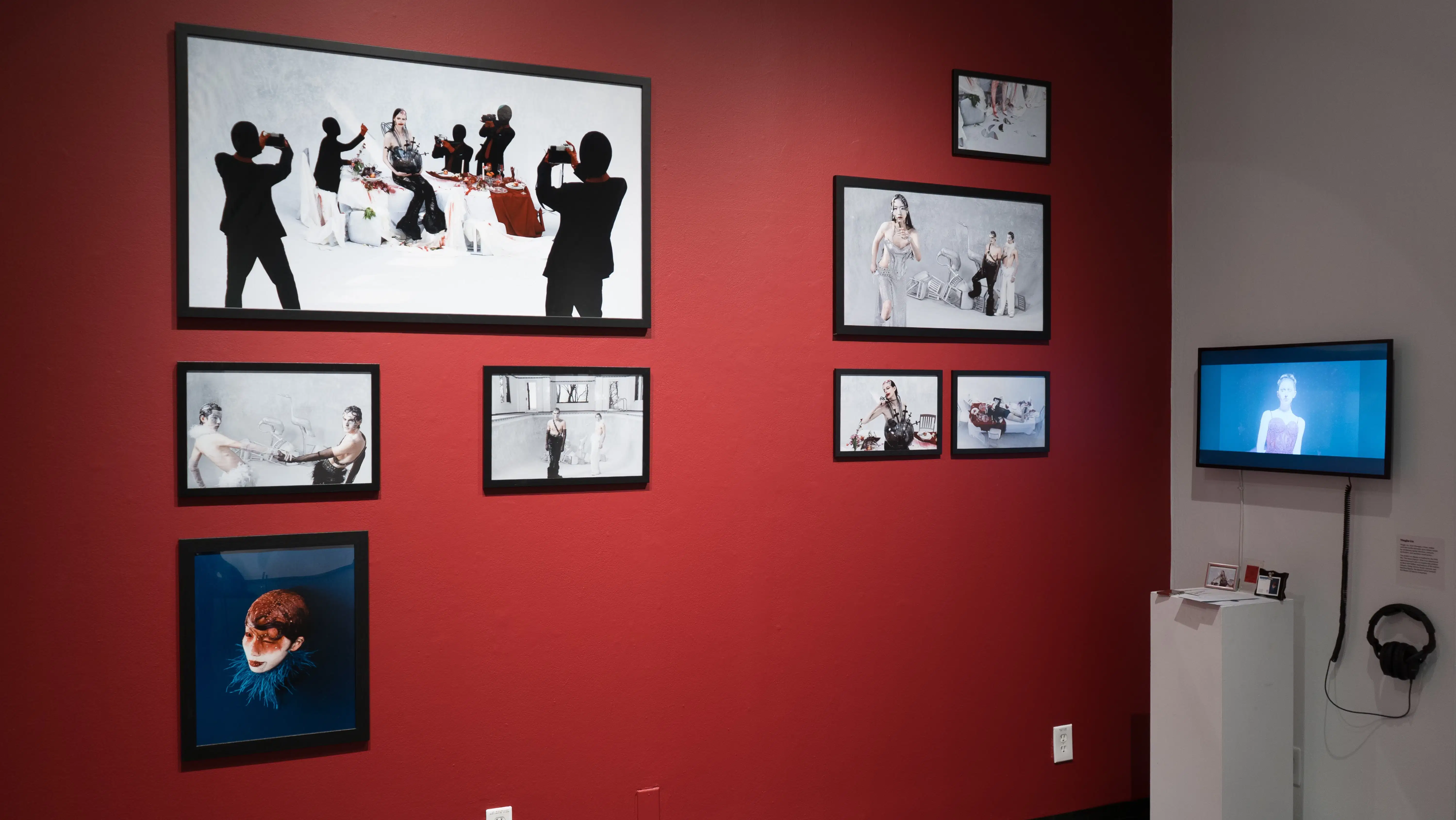 Black framed images hanging on a red wall in the SVA Gramercy Gallery. A screen hangs on a white wall perpendicular to them.