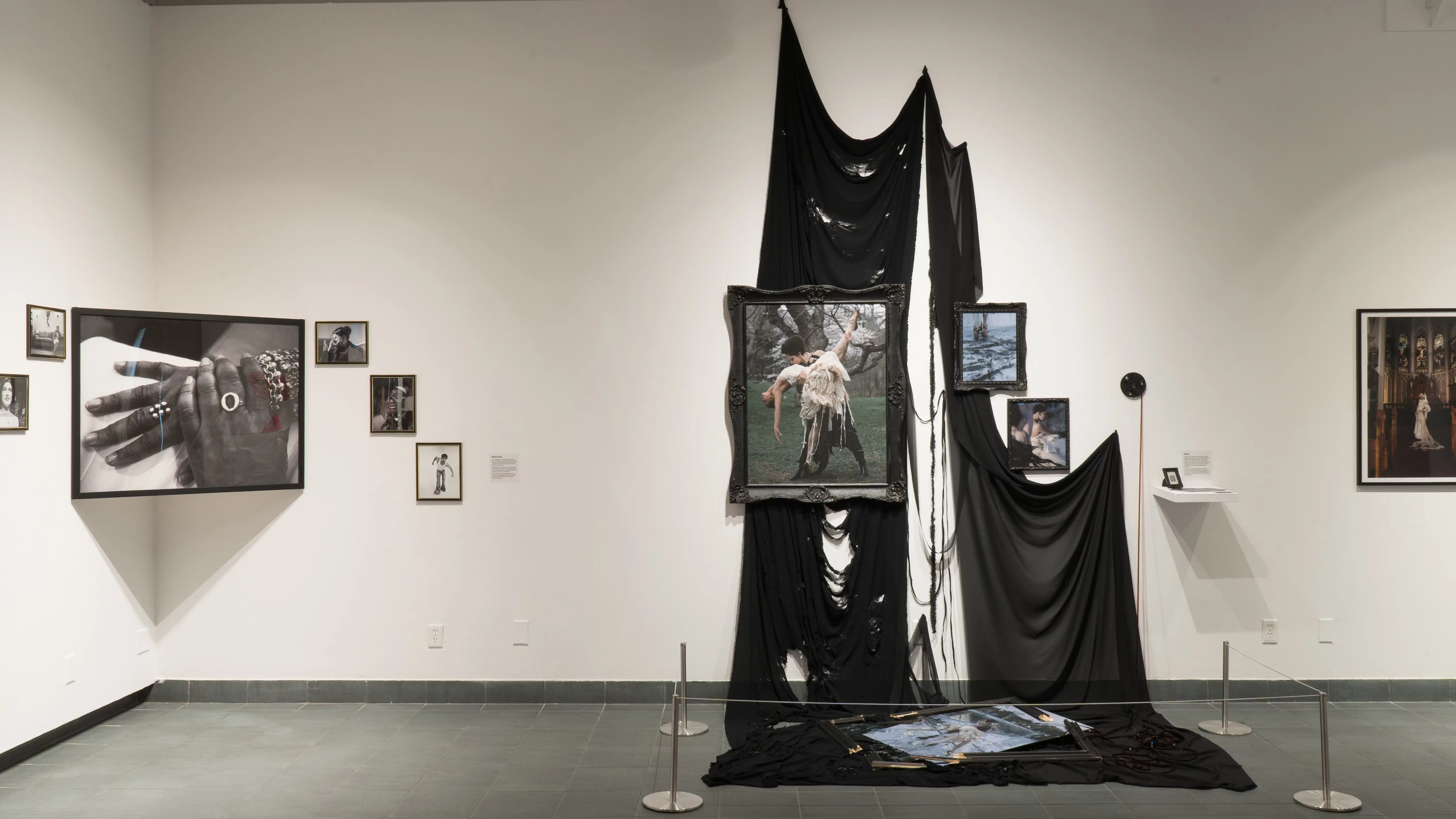 Black and white pictures hang in the corner of the gallery, and to their left is a gothic-looking display. Images hang in elaborate frames, while black fabric drapes behind them, one frame and picture lies shattered on the cloth-covered floor.