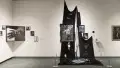 Black and white pictures hang in the corner of the gallery, and to their left is a gothic-looking display. Images hang in elaborate frames, while black fabric drapes behind them, one frame and picture lies shattered on the cloth-covered floor.