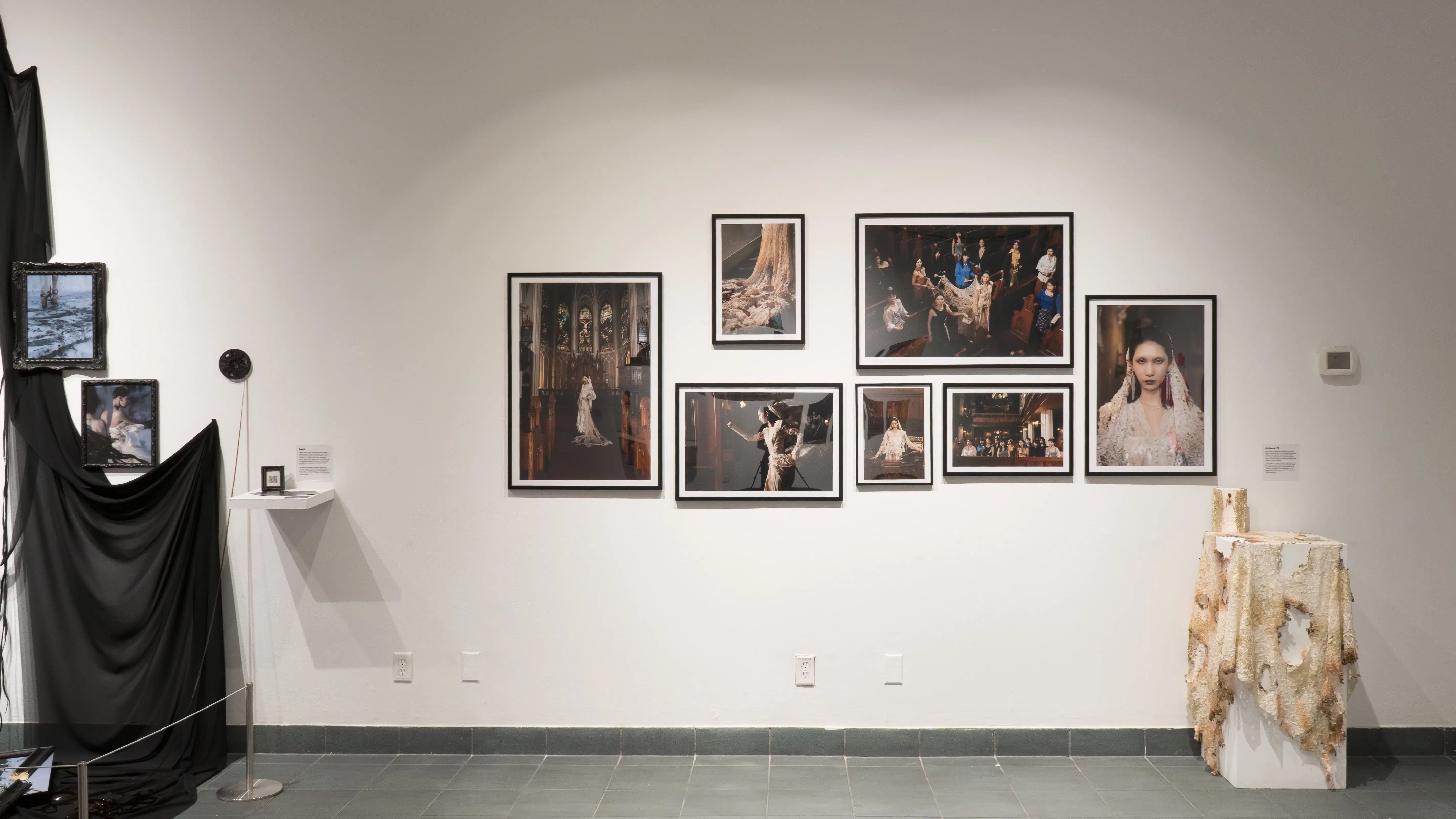 Various images of a model in a church hang on a wall in the gallery. Below and to the right of these images is a pedestal draped in burnt lace.