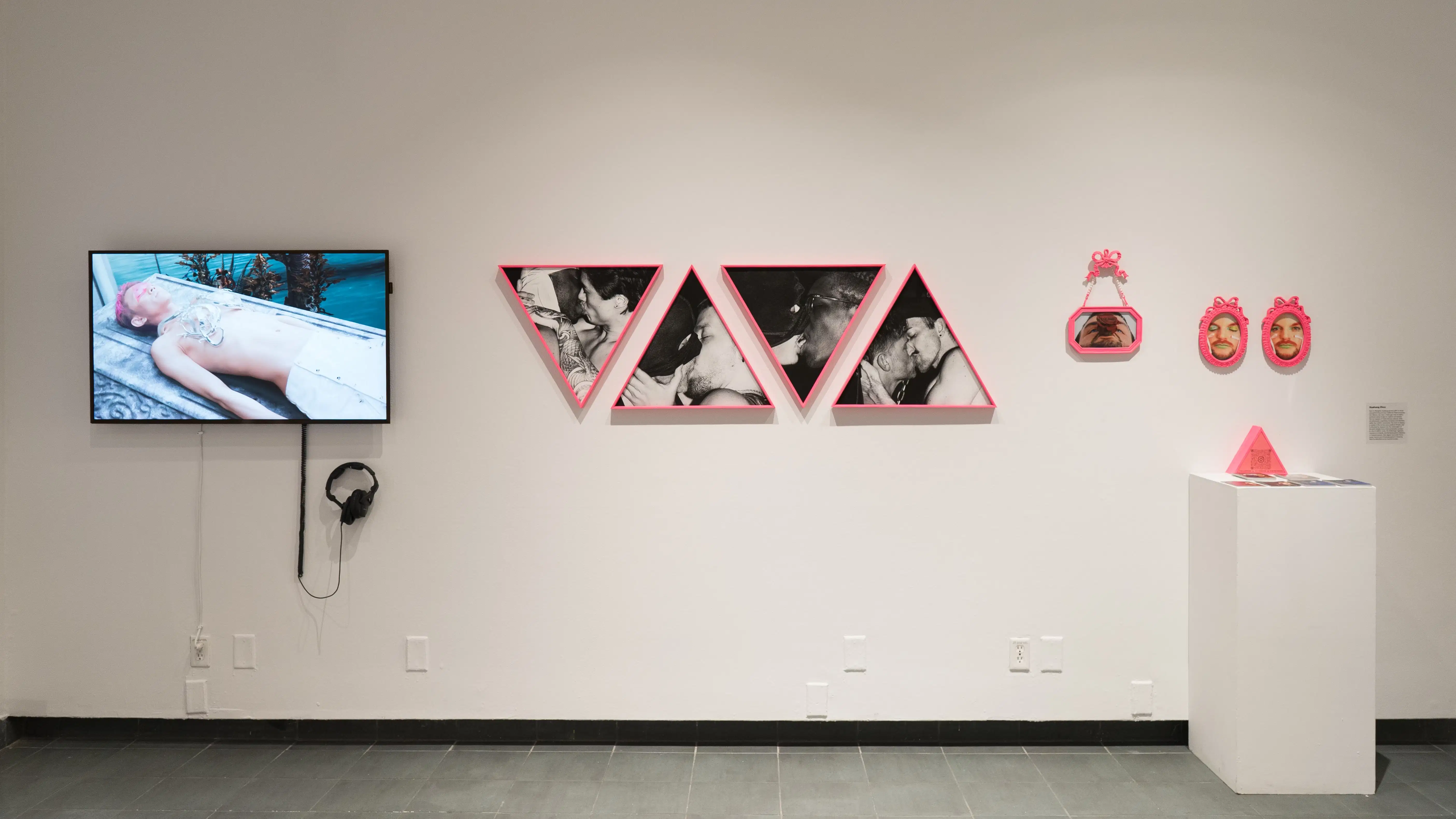 On a wall in the SVA Gramercy Gallery, video art is displayed on a monitor. To the right of that there are images of men kissing hang in pink, triangular frames, as well as smaller frames of a model smirking at the audience.