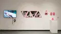 On a wall in the SVA Gramercy Gallery, video art is displayed on a monitor. To the right of that there are images of men kissing hang in pink, triangular frames, as well as smaller frames of a model smirking at the audience.