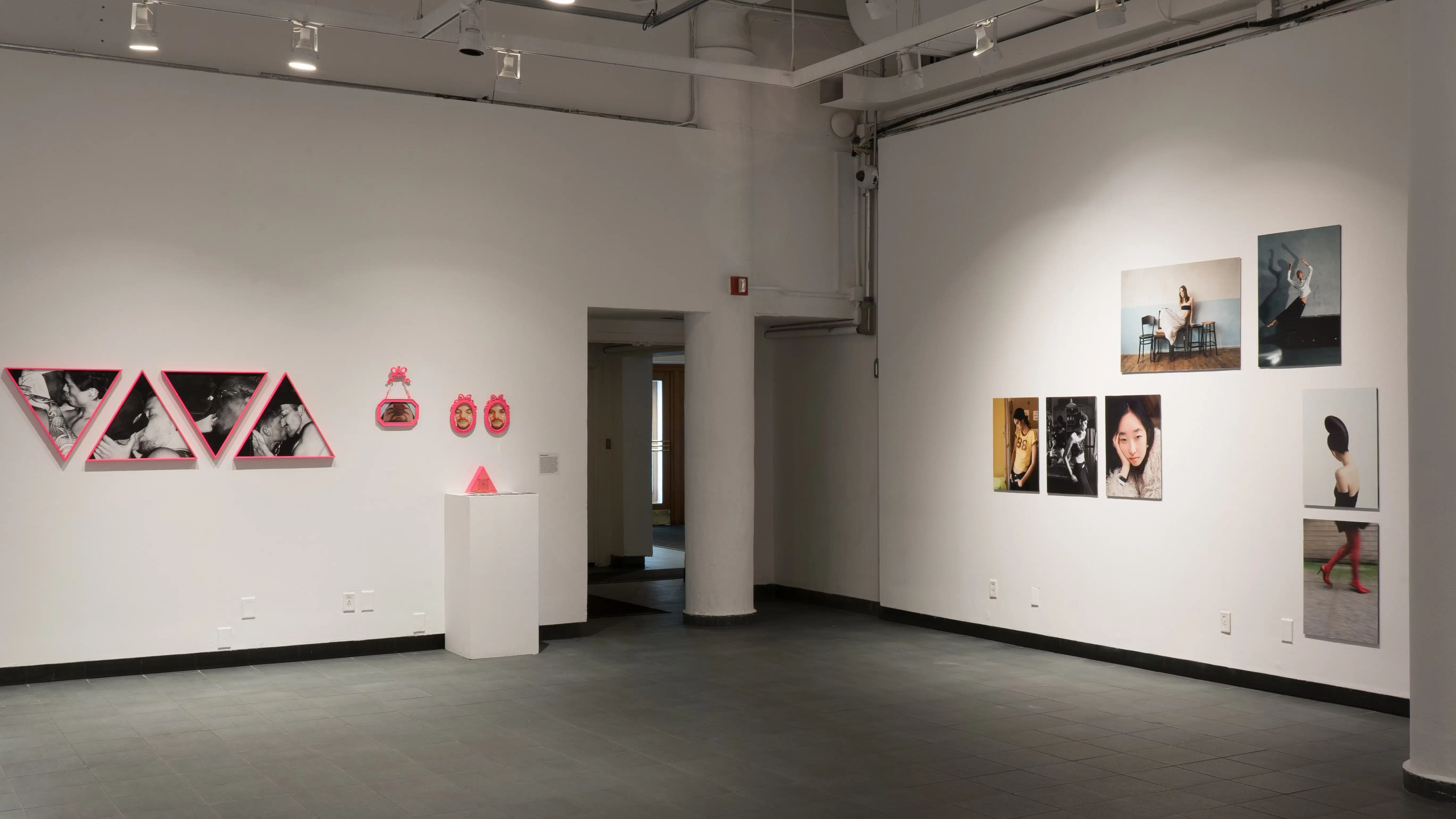 In the gallery, student photography hangs on the wall. On one wall, the works are in bright pink frames, and a pedestal holds a triangular pink sculpture. The works perpendicular to those aren’t framed, and feature images of models in various poses.