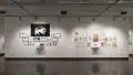 A view of the east wall at the SVA Chelsea Gallery. On the left is a black and white project hung salon style. There is a large black flag with a cat with a sword in white at the top, under the words, “Bonny sets sail.” Another project hung salon-style is on the right, with many colorful framed prints under the title, “For All We Know.”