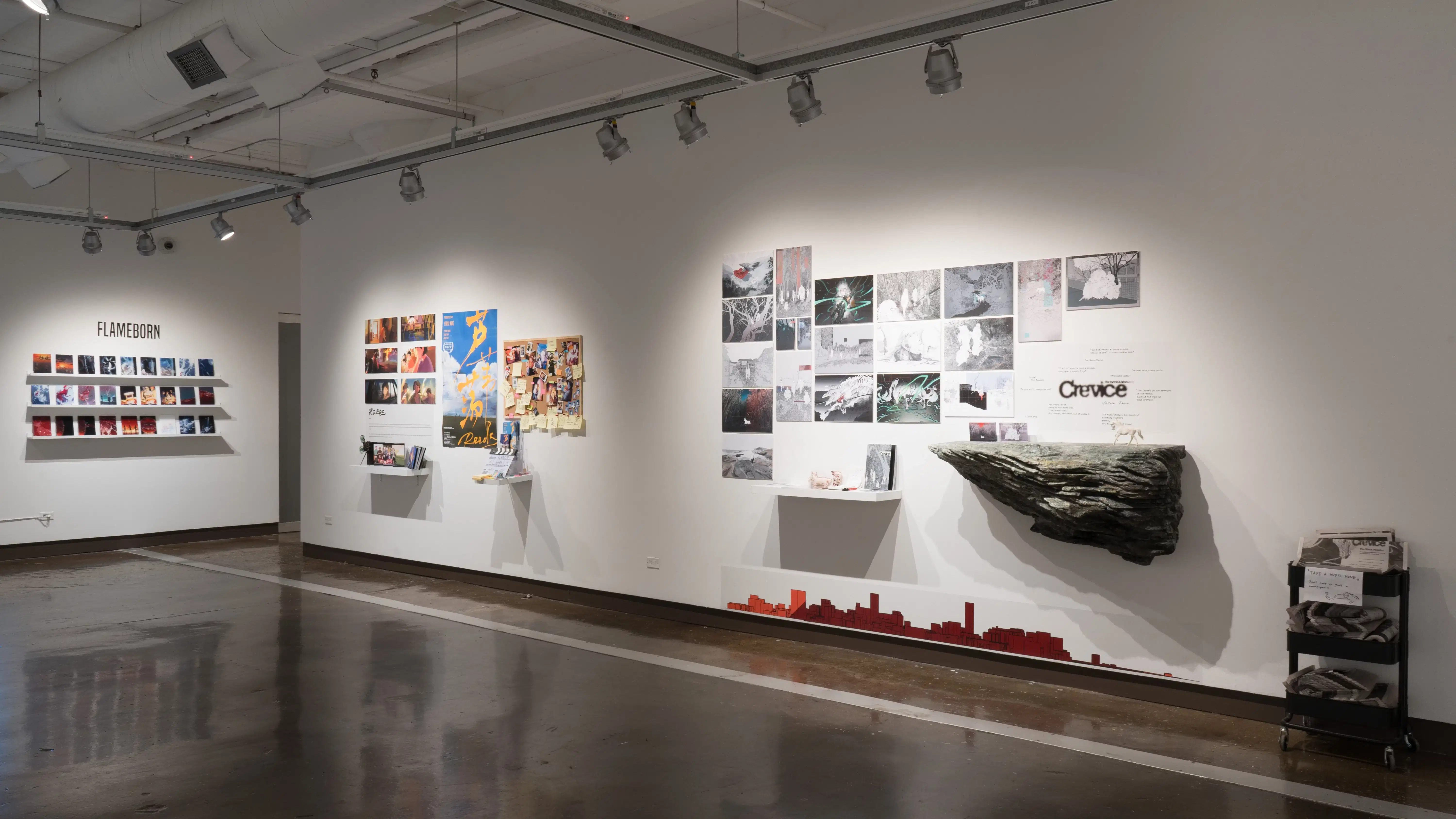 An angled view of the south wall in Gallery 2 shows a project with a large imitation rock on the wall below the title “Crevice.” There are many largely black and white prints hung above it. The the left theres a pin board with postit notes and small cards, and many colorful prints next to it.