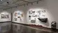 An angled view of the south wall in Gallery 2 shows a project with a large imitation rock on the wall below the title “Crevice.” There are many largely black and white prints hung above it. The the left theres a pin board with postit notes and small cards, and many colorful prints next to it.