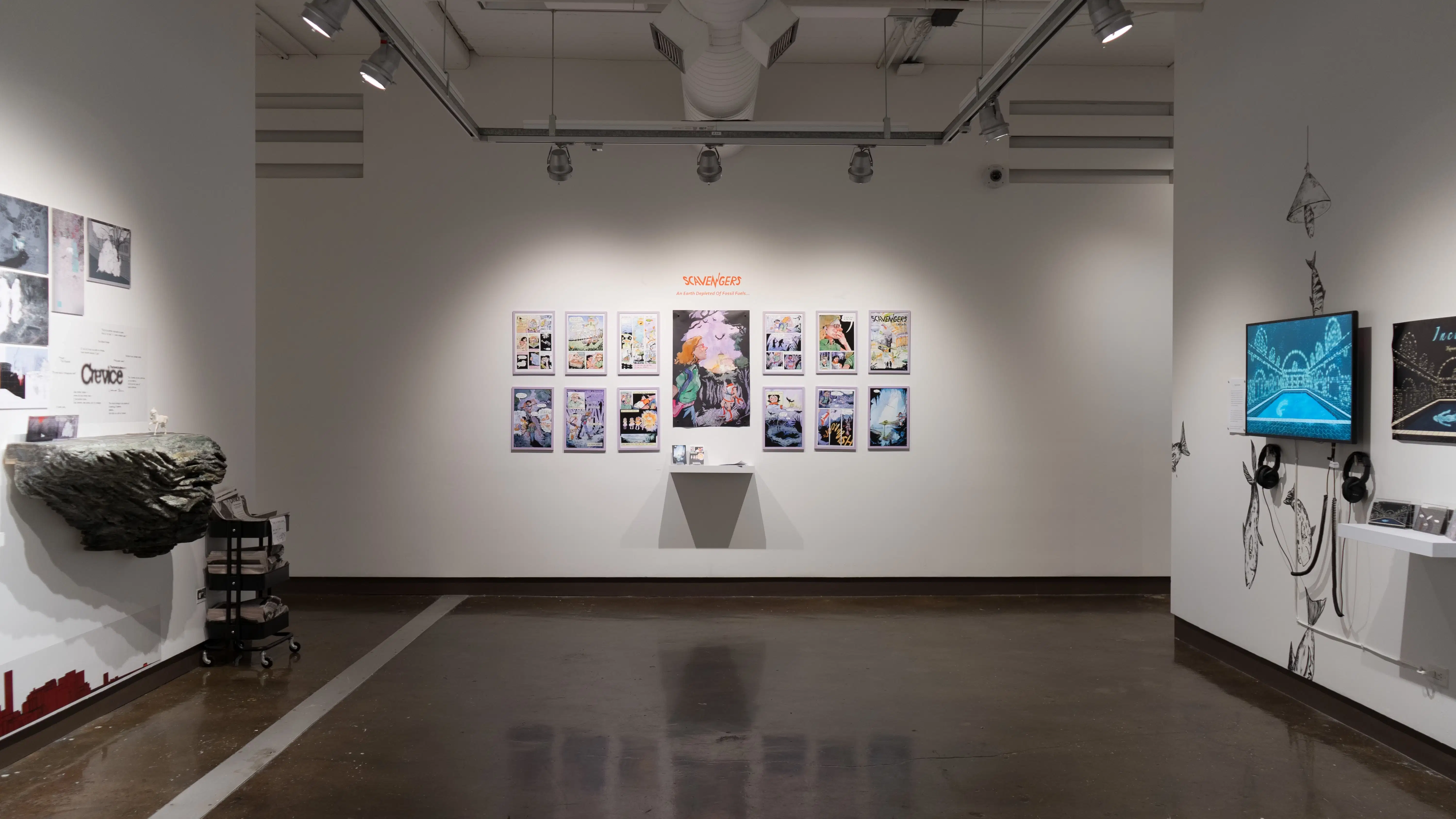 Three illustration projects are visible on one side of gallery two. On the far wall, a large, centered poster is flanked on both sides by two rows of framed prints. To the left, a large faux rock juts out of the wall below several prints pinned to the wall. To the right, a monitor and headphones are surrounded by fish painted in black on the wall.